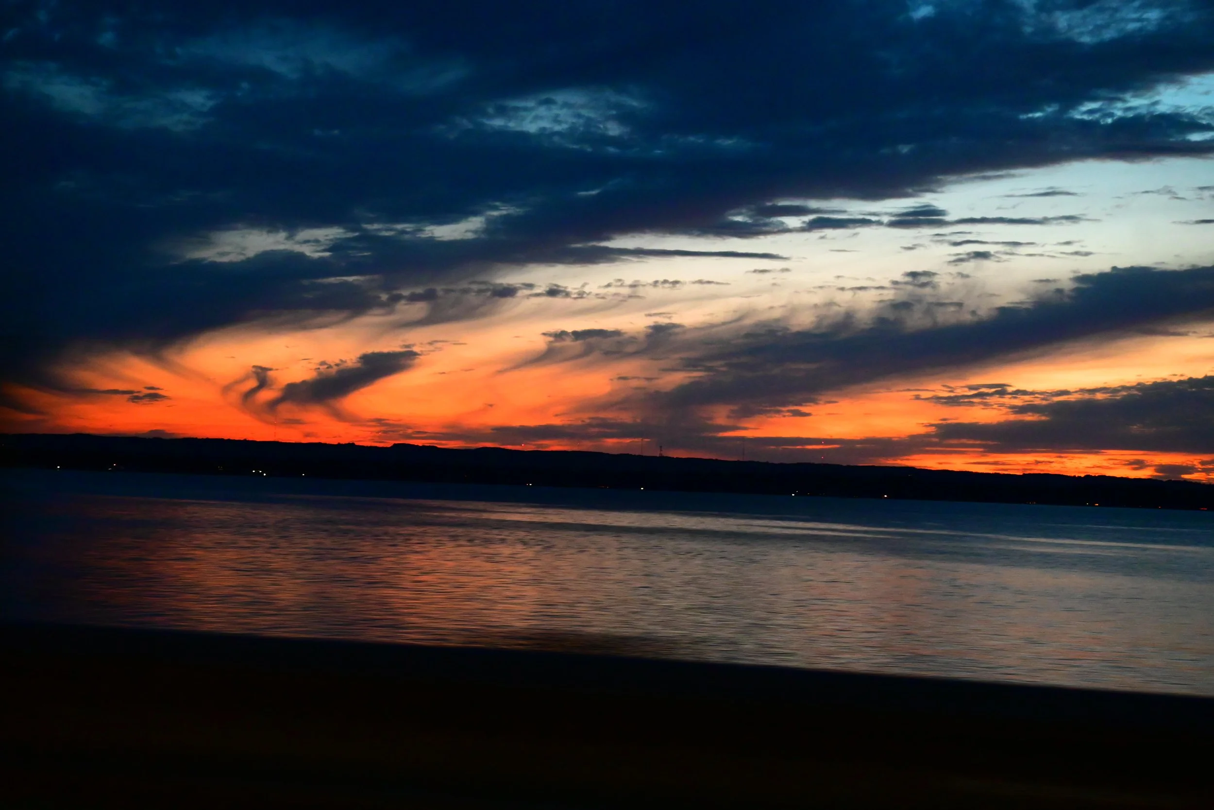 A sunset over a body of water with a dark silhouetted shoreline, featuring dark clouds in the sky with hints of orange and yellow hues.
