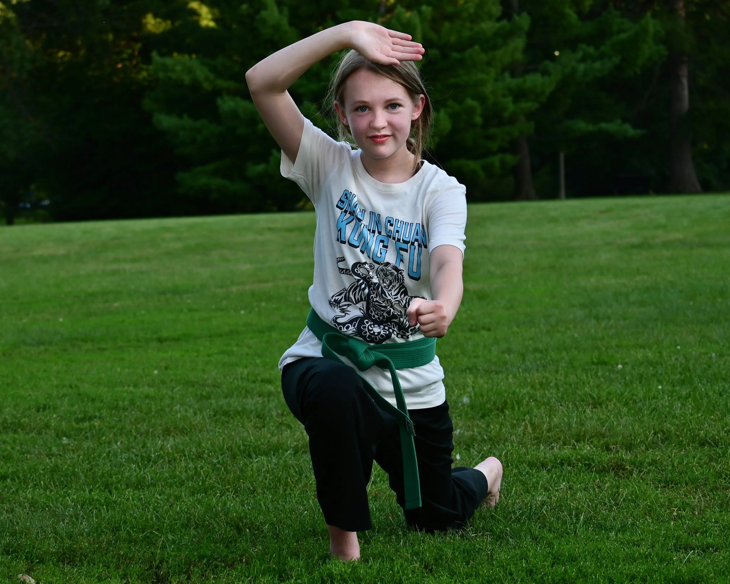 A young girl practicing martial arts outdoors on a grassy field, wearing a martial arts uniform with a green belt, in a pose with one arm extended and the other raised above her forehead, with trees in the background.