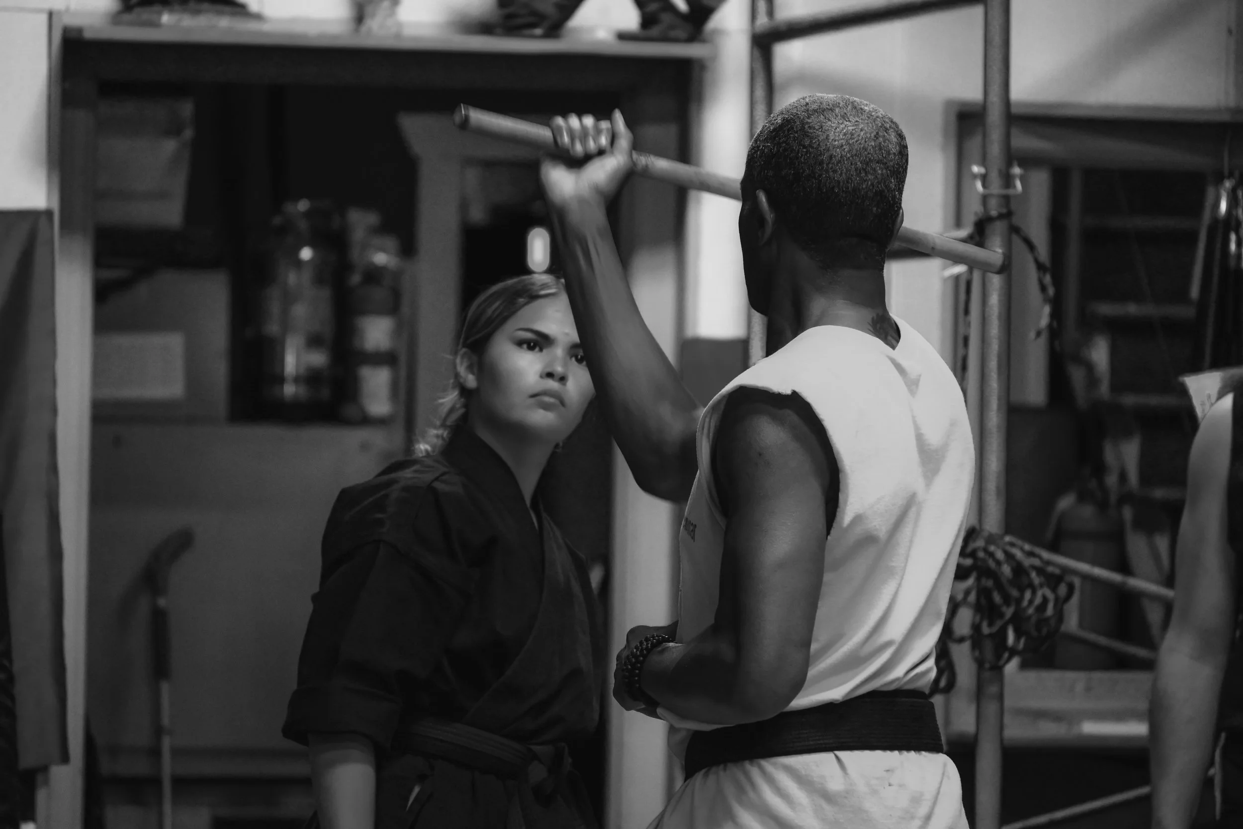 A woman in martial arts attire practicing a weapon skill with a staff as a man in a sleeveless shirt assists her, standing in a room with shelves and equipment.