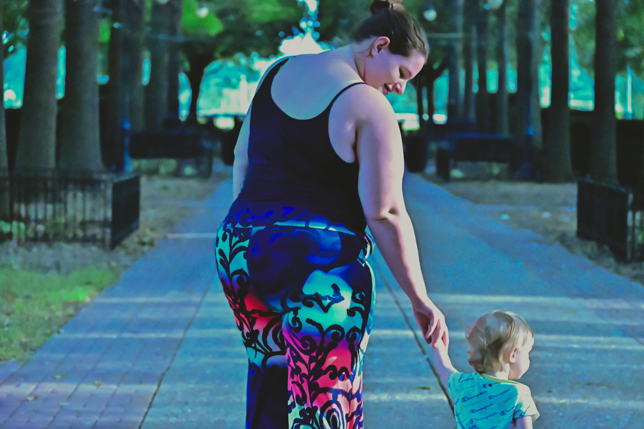 A woman with dark hair tied back, wearing a black tank top and vibrant, patterned leggings, is walking a young boy with blond hair who is wearing a blue shirt with a pattern. They are outdoors in a park with a paved path, trees, benches, and greenery in the background.