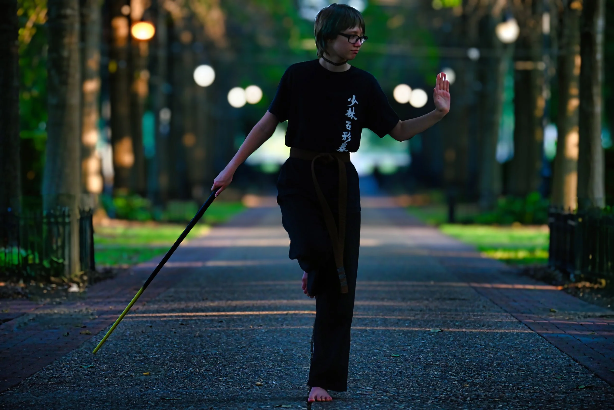 A young person with short hair, glasses, and a choker walks barefoot on a paved pathway through a lush green park at dusk, holding a walking stick in one hand and raising the other hand in a shushing gesture.