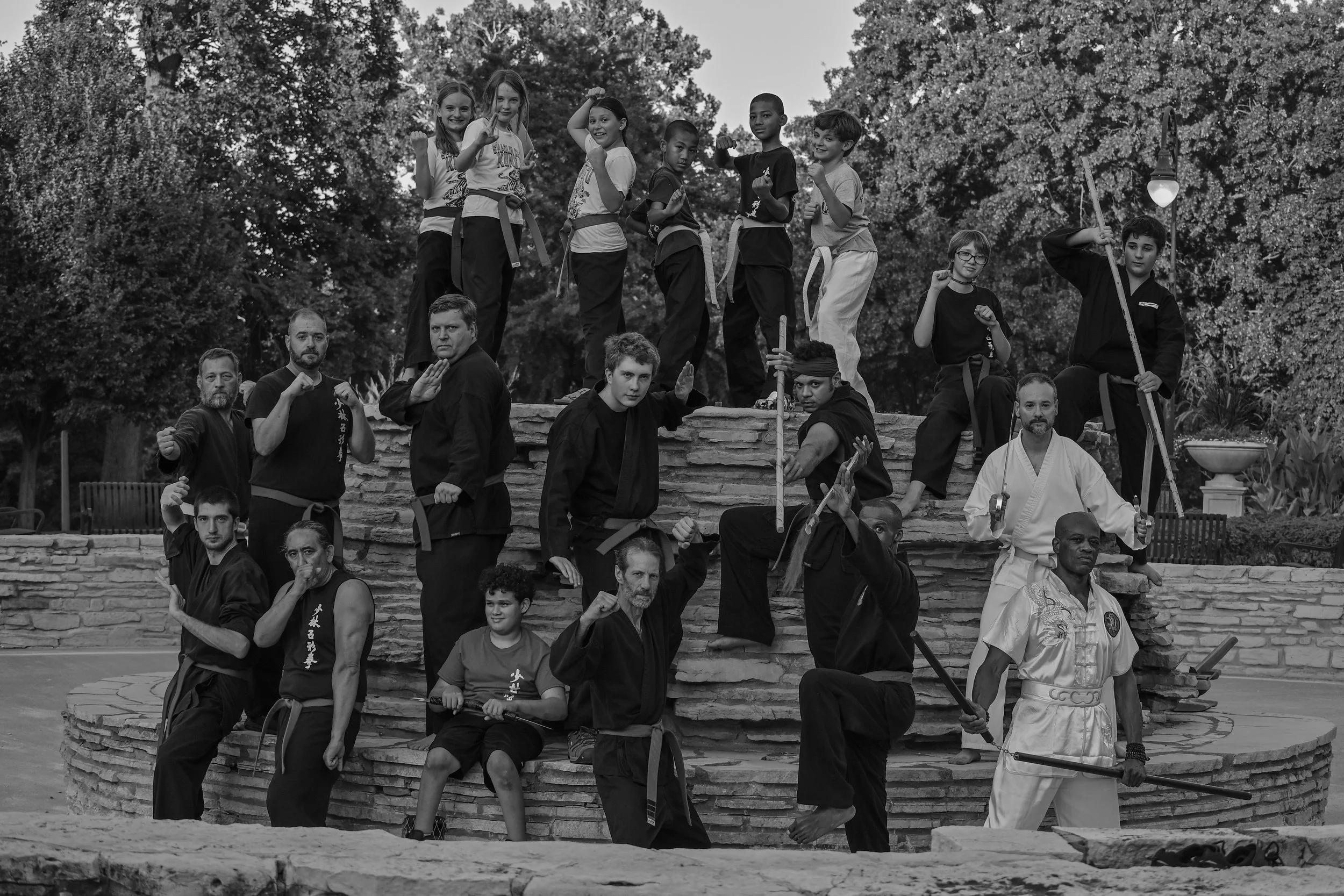 A group of martial arts students and instructors in uniform posing outdoors, many flexing and making fists, with trees in the background.