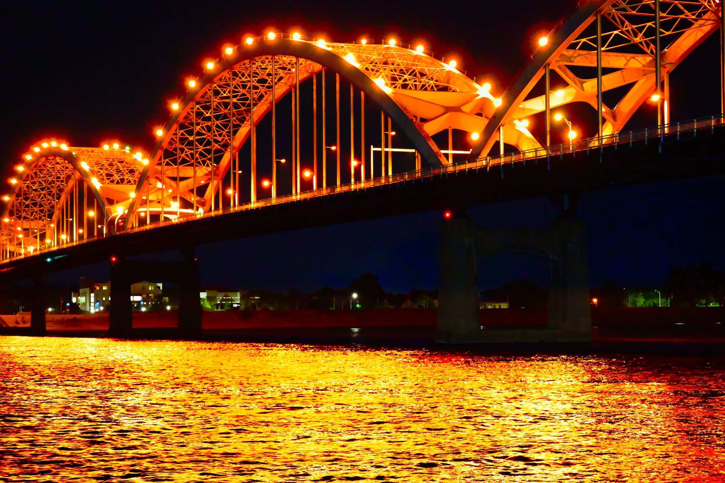 Nighttime view of a brightly lit suspension bridge over a body of water, with reflections shimmering on the surface.