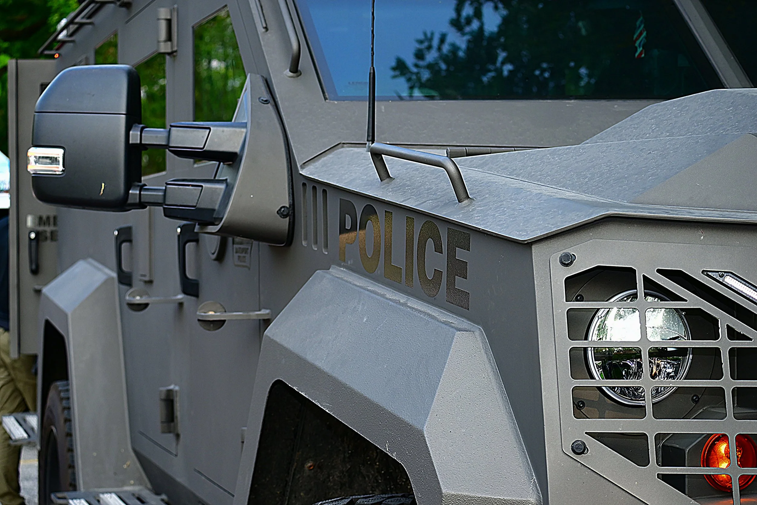 Close-up of a police armored vehicle with a gray metal exterior, black side mirror, and reflectively tinted window, parked outdoors with green trees in the background.