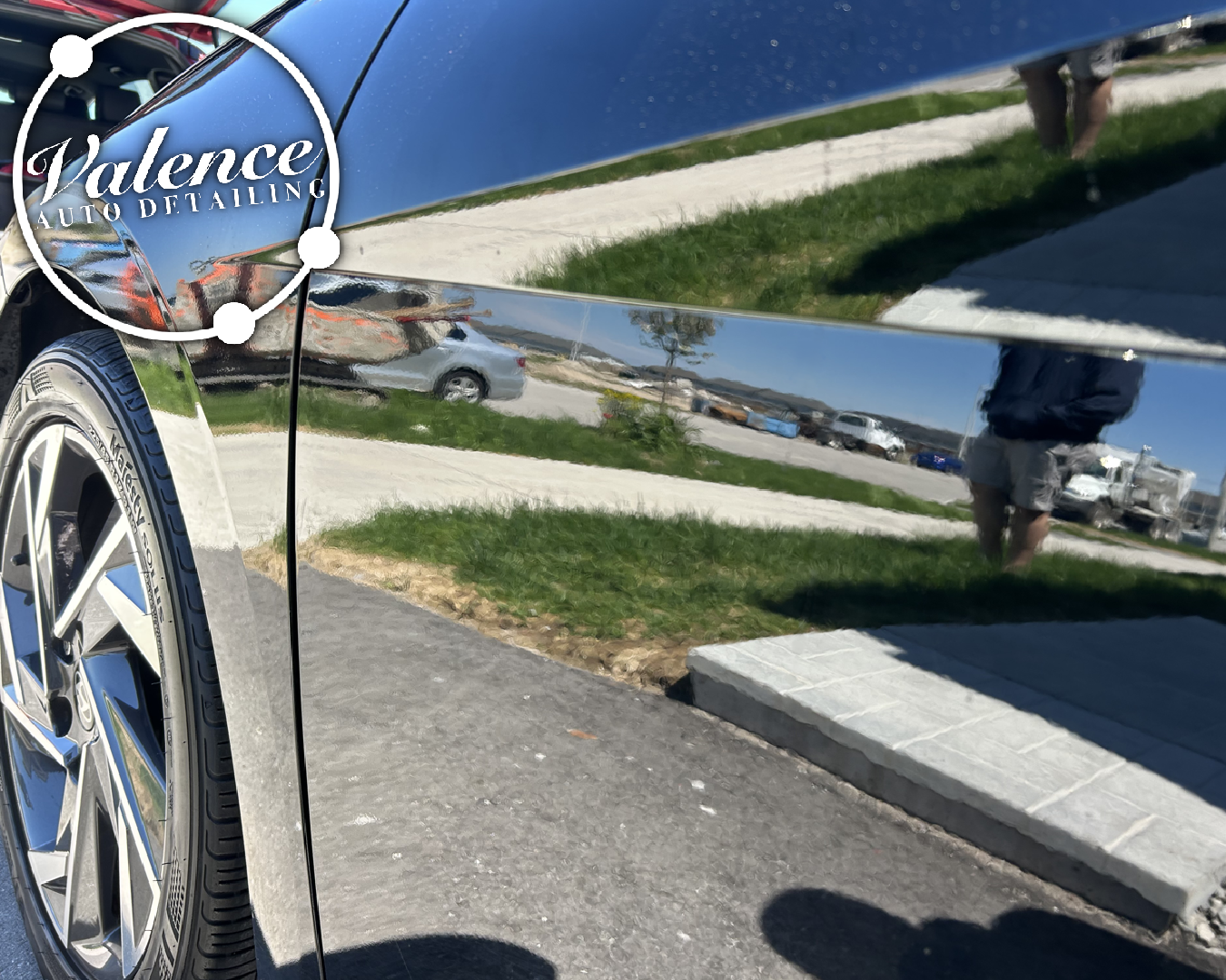 Close-up of a shiny black car with a reflection of a parking lot, grass, sidewalk, and a person wearing shorts taking the photo.