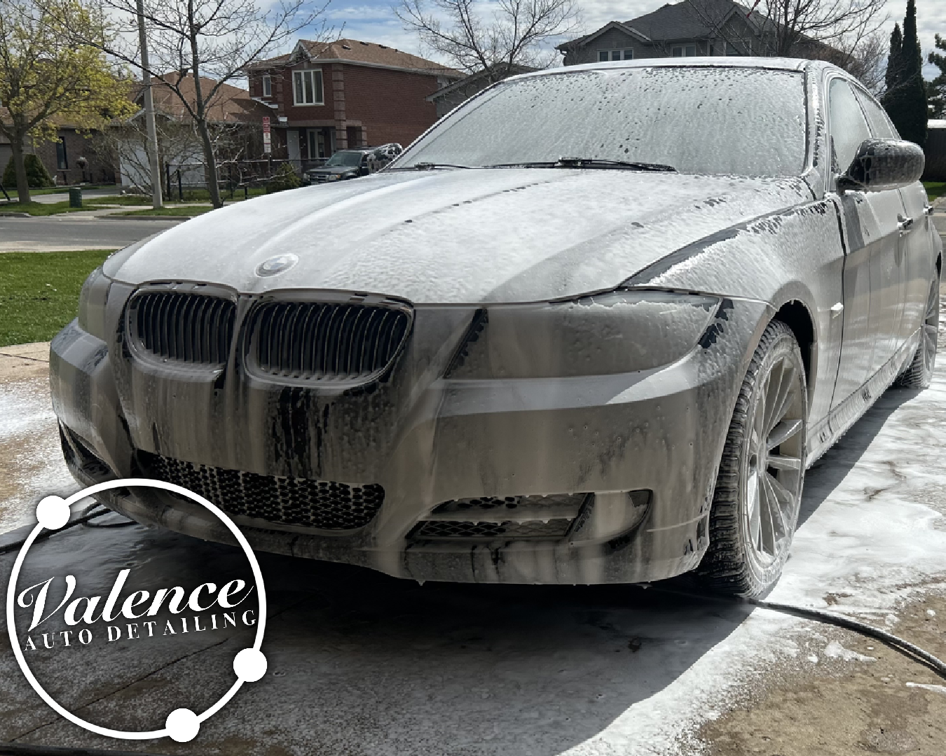 A silver BMW vehicle covered in soap and foam during a car wash at Valence Auto Detailing, parked on a driveway with houses and trees in the background.
