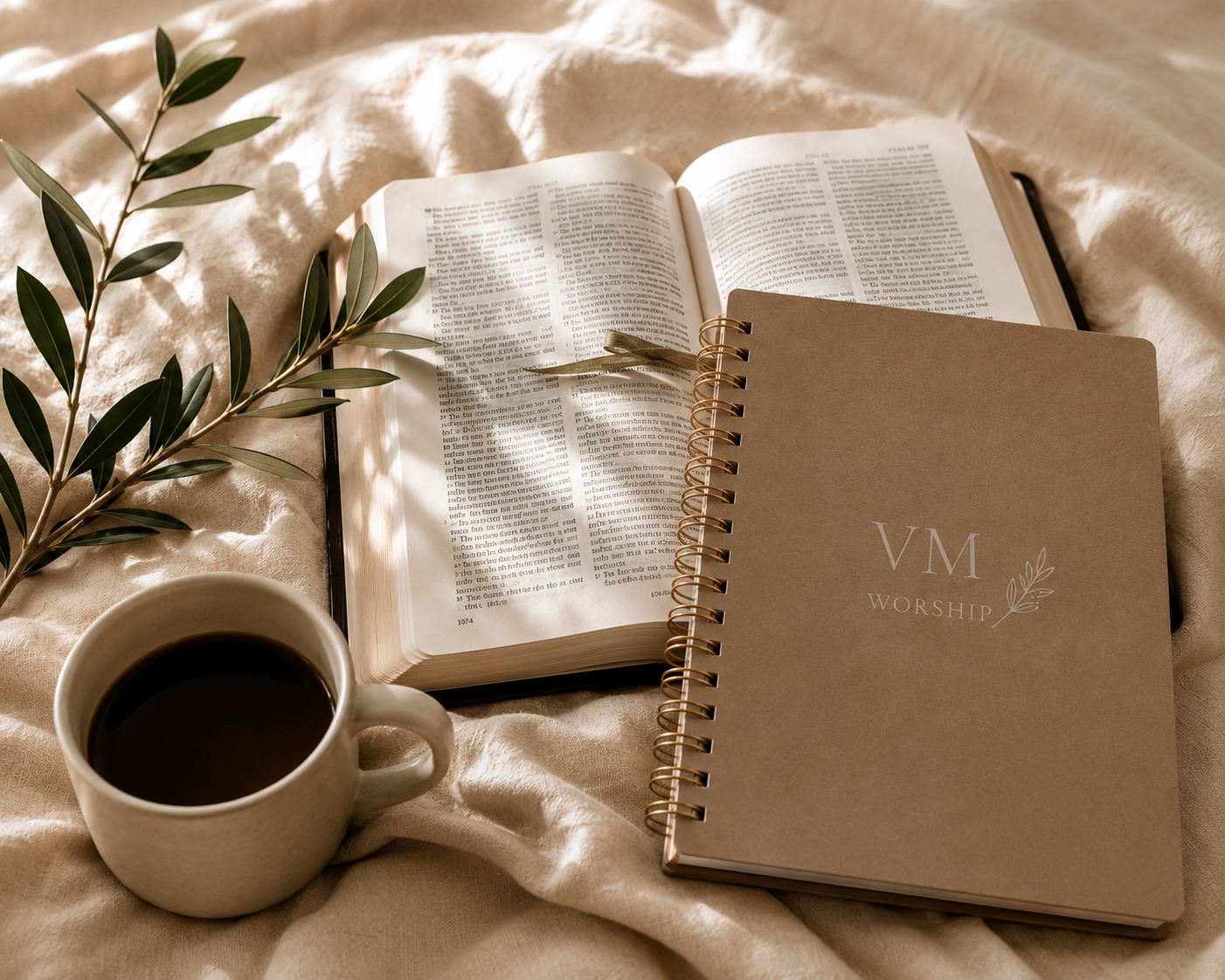 An open Bible, a closed worship planner labeled "VM Worship," a coffee mug filled with black coffee, and a green leafy branch, all arranged on a soft beige fabric surface.