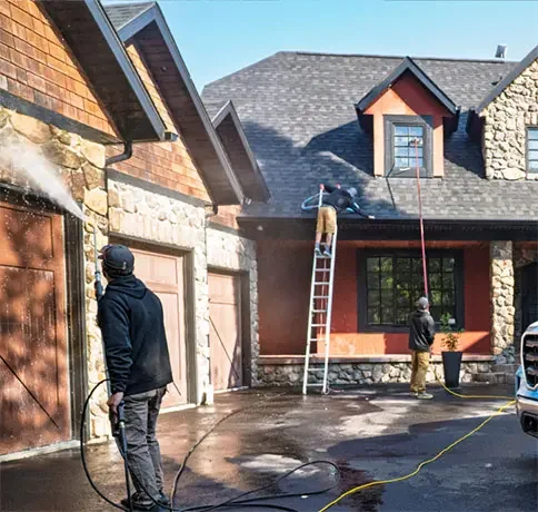 Two people power washing a residential house exterior with a ladder leaning against the house and a car parked nearby.