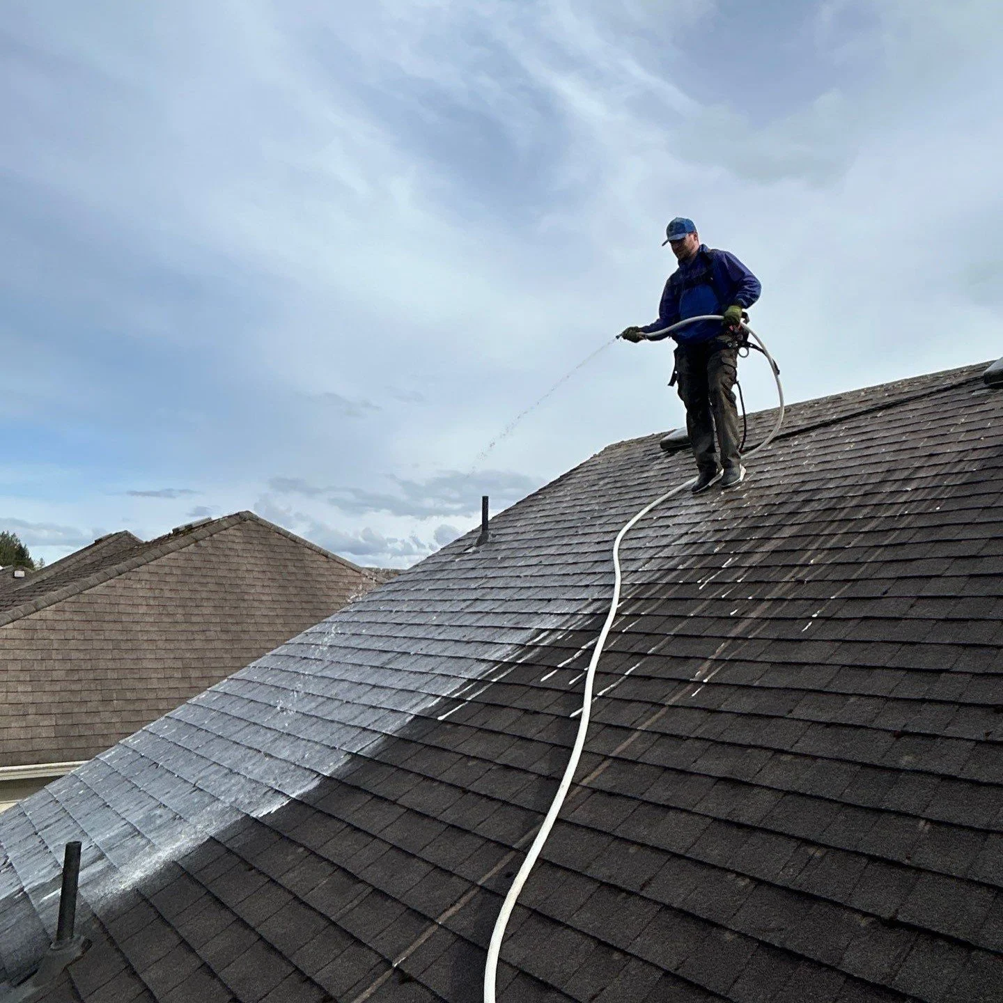 A worker on a roof power washing shingles with a spray wand, water running down the surface, under a cloudy sky.