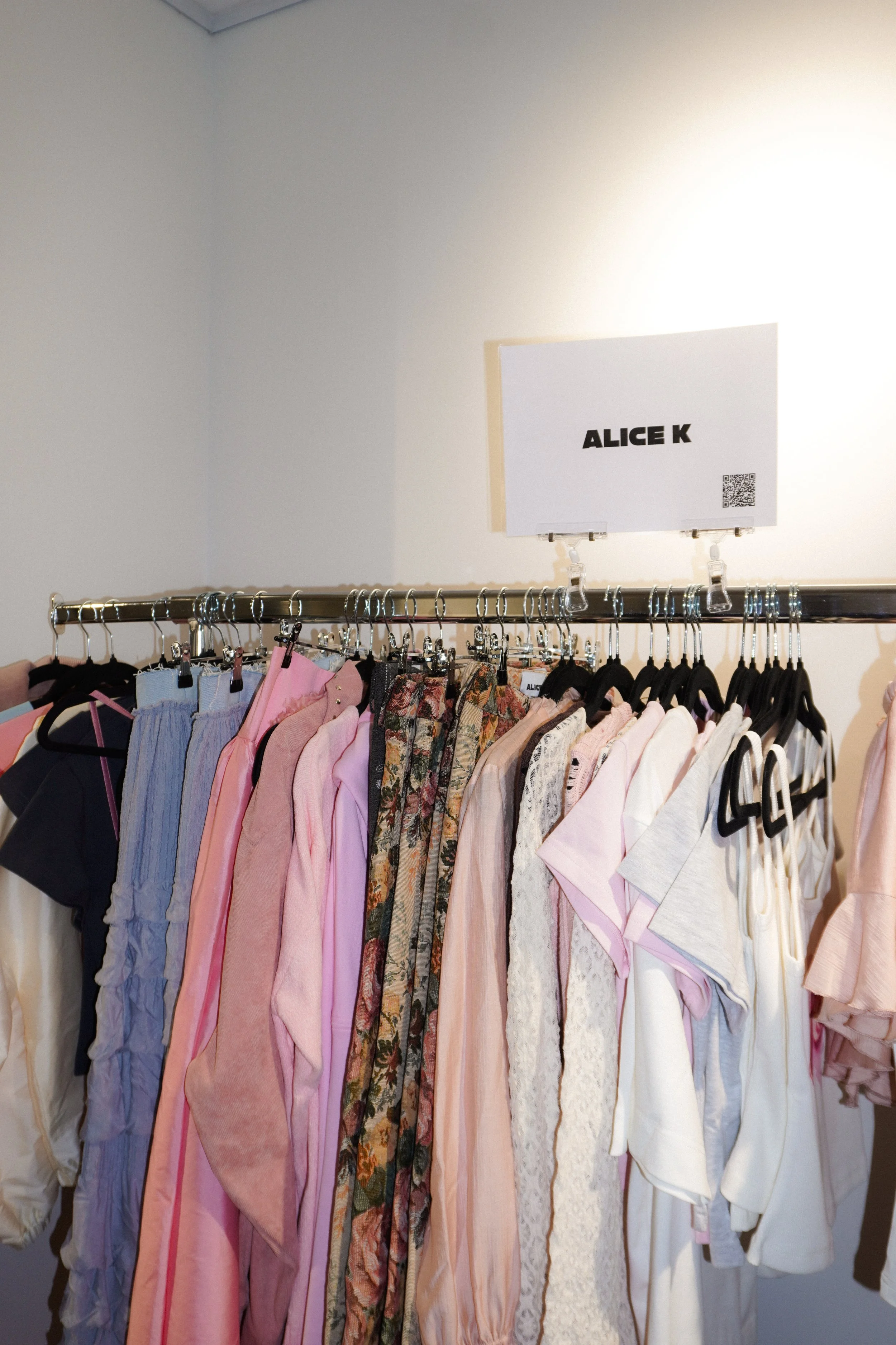 Clothing rack with various women's pajamas and sleepwear in pink, black, floral, white, and beige, organized on hangers beneath a sign with 'ALICE K' and a QR code.