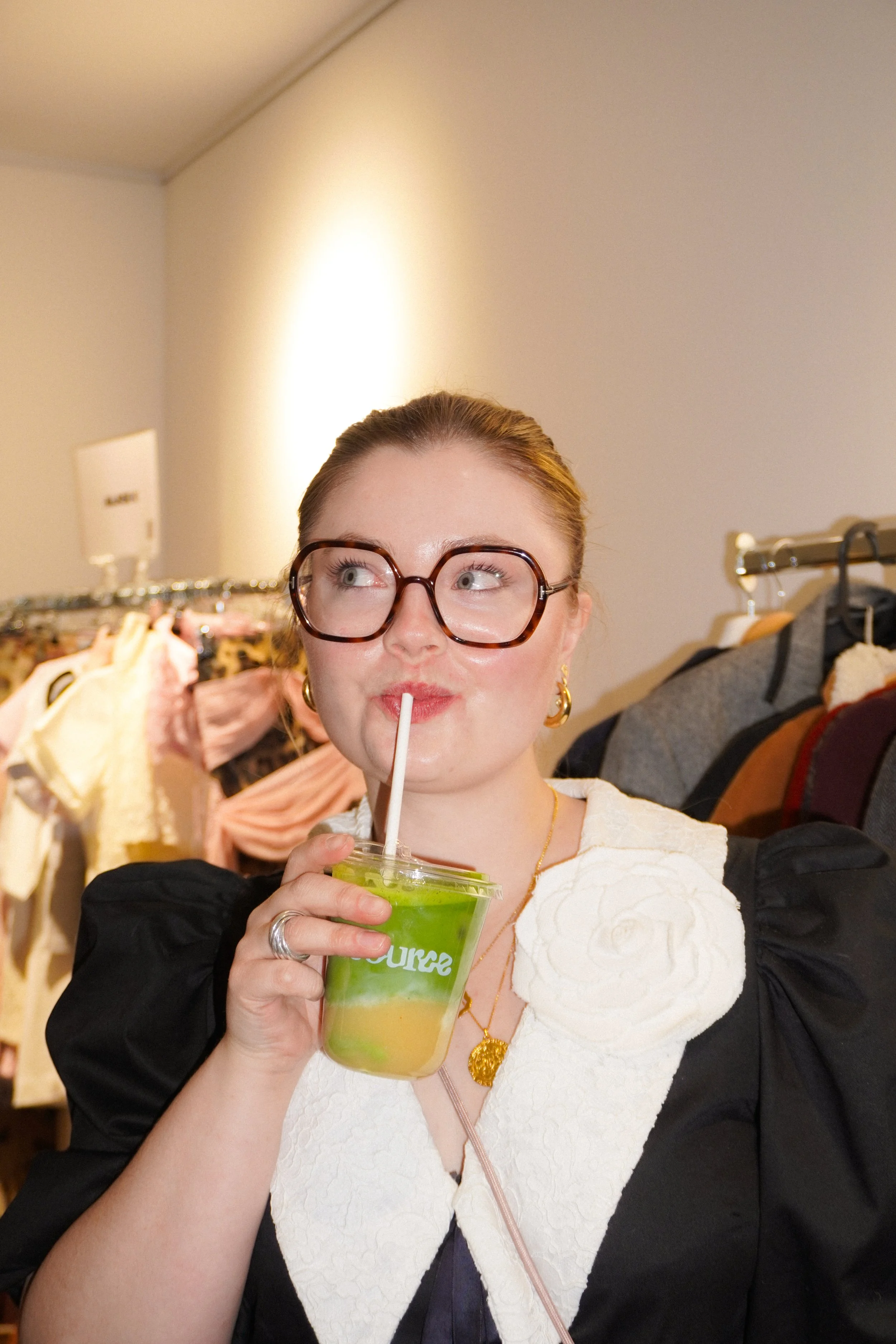 A woman with glasses drinking a green and yellow beverage with a straw. She is wearing a white top with black sleeves, gold jewelry, and is standing in a clothing store.