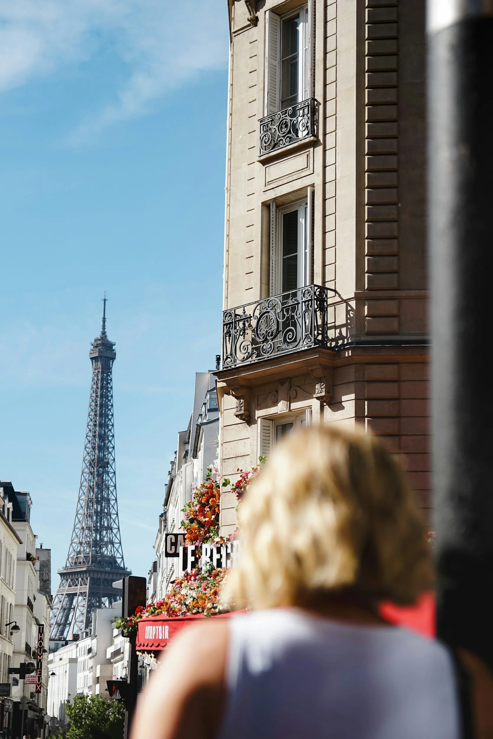 Street scene in Paris with the Eiffel Tower in the background, showing buildings with French architecture, balconies, flower decorations, and some signage, view partially obstructed by a person in the foreground.