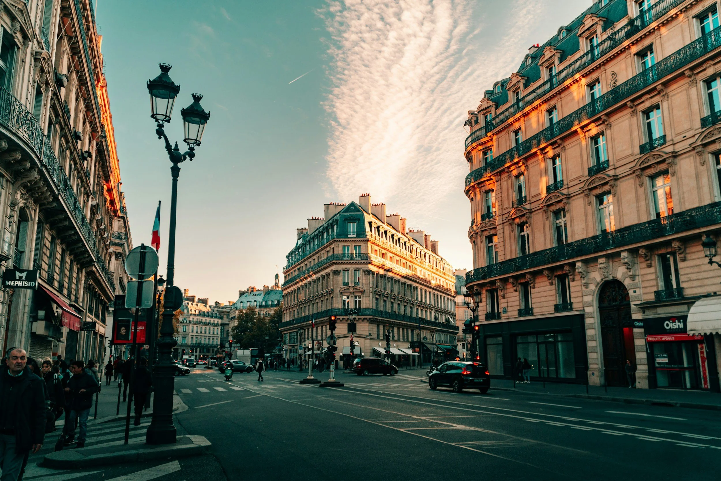 City street scene in Paris with classic Parisian architecture, ornate buildings, and a clear sky with clouds. People are walking on the sidewalk, and cars are on the road. A street lamp is visible in the foreground.