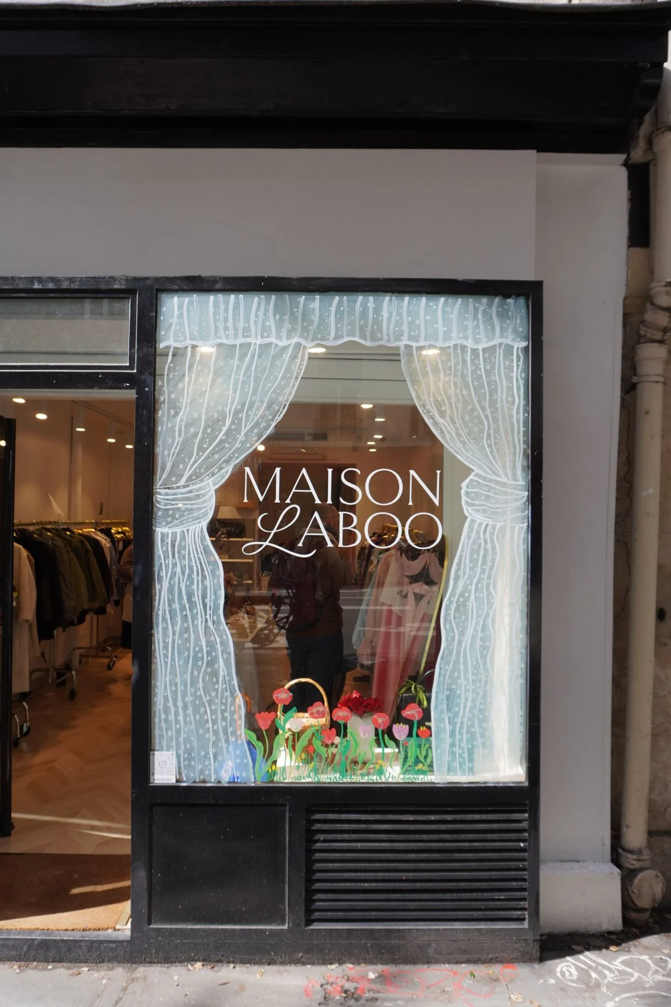 storefront window with white curtains and the words 'MAISON LABO' was visible inside the store. The display has colorful flowers at the bottom of the window.