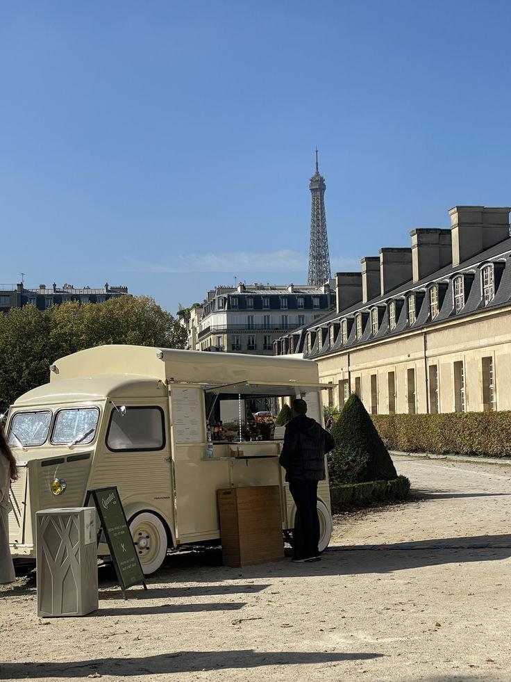 A small cream-colored mobile food truck parked on a dirt path in front of classic Parisian buildings with the Eiffel Tower visible in the background. A person is standing at the truck, ordering or paying.
