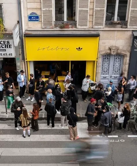 A group of people gathered outside a storefront with a yellow awning that has the word "solebox" and the Adidas logo. Some are socializing, and a person is playing guitar.