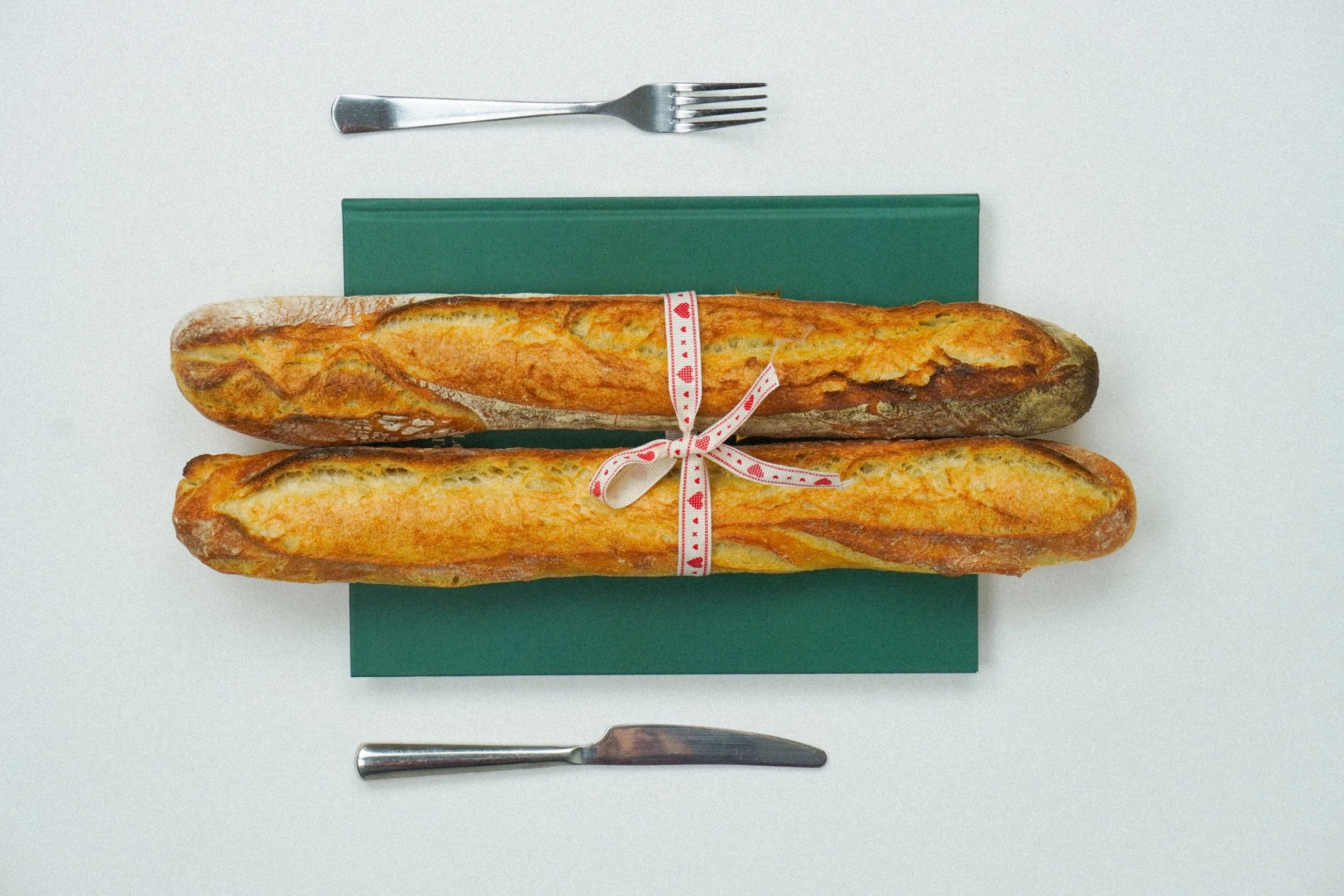Two baguettes tied with a white ribbon with red hearts, placed on a green cutting board, with a silver fork above and a silver knife below, all on a white background.
