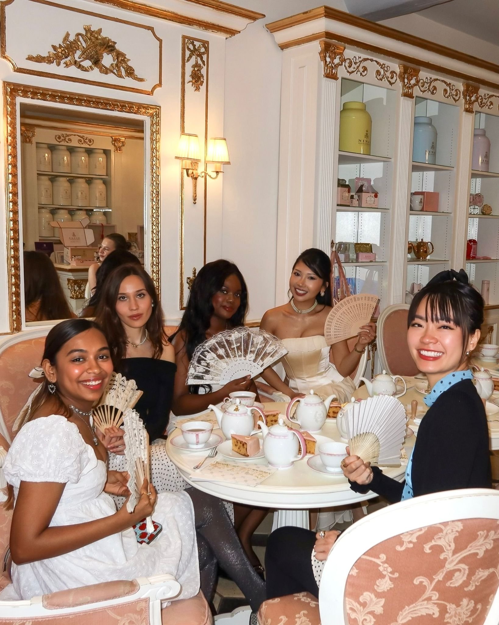 A group of women sitting around a table during afternoon tea at an elegant, decorated venue. They are smiling, holding fans, and dressed in sophisticated attire. The table is set with teapots, cups, slices of cake, and other tea-time items, and the background features white and gold ornate decor with shelves displaying teapots and decorative items.