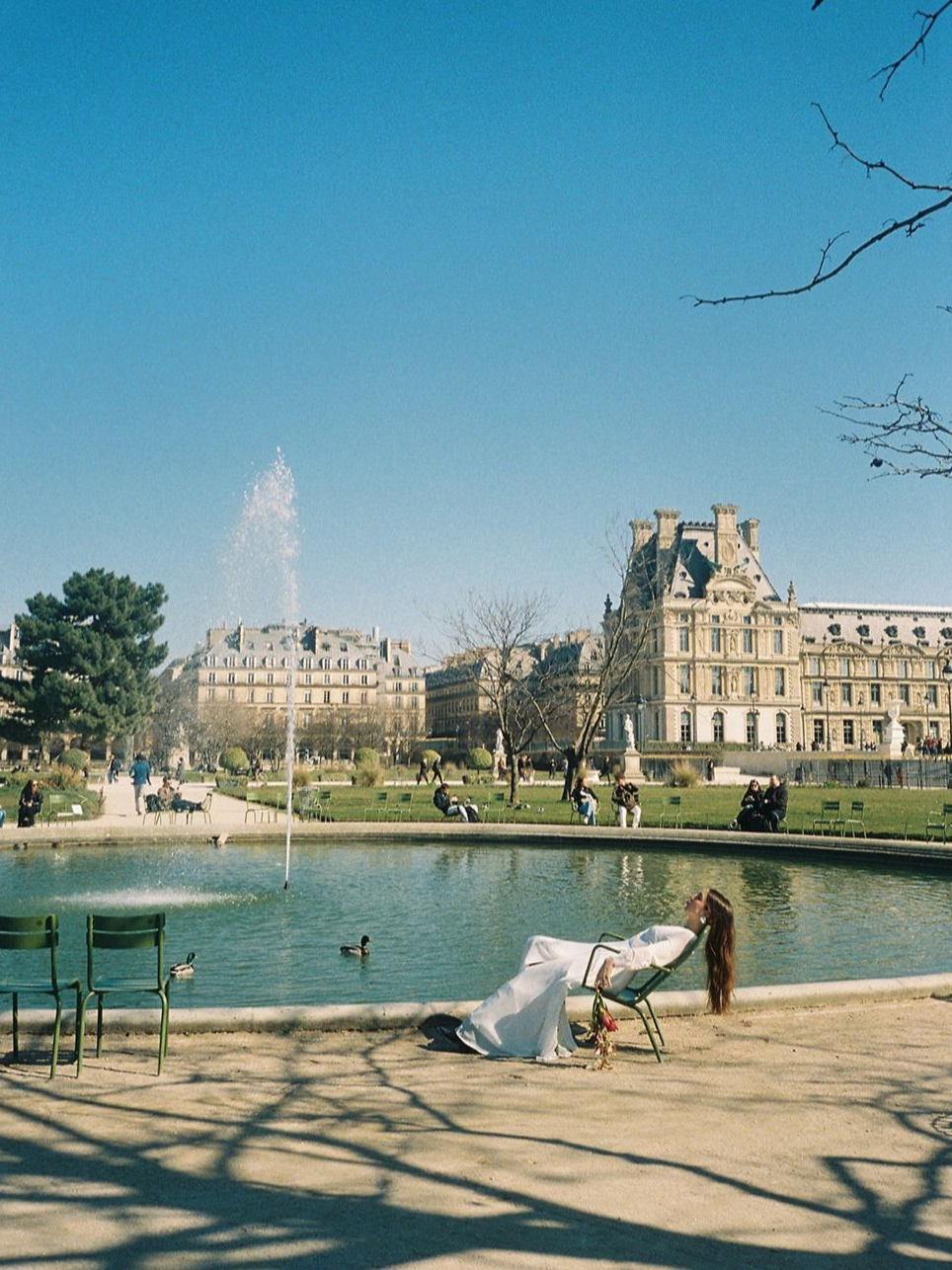 A woman in a white dress lies on a lounge chair by a fountain in a park, with historic buildings and people sitting on benches in the background under a clear blue sky.