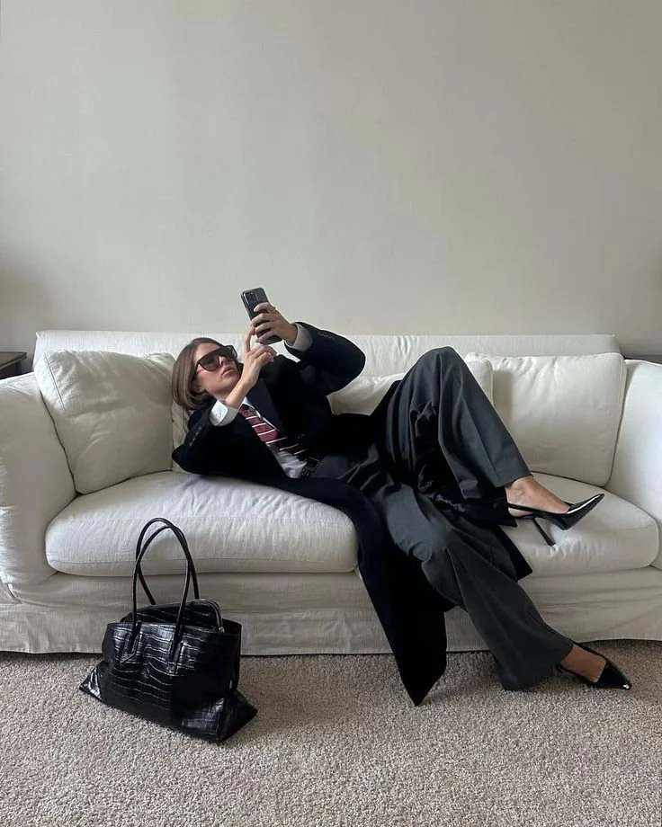 A woman in a black suit and sunglasses is reclining on a white couch, taking a selfie with her phone. She has a black handbag on the carpeted floor next to the couch.