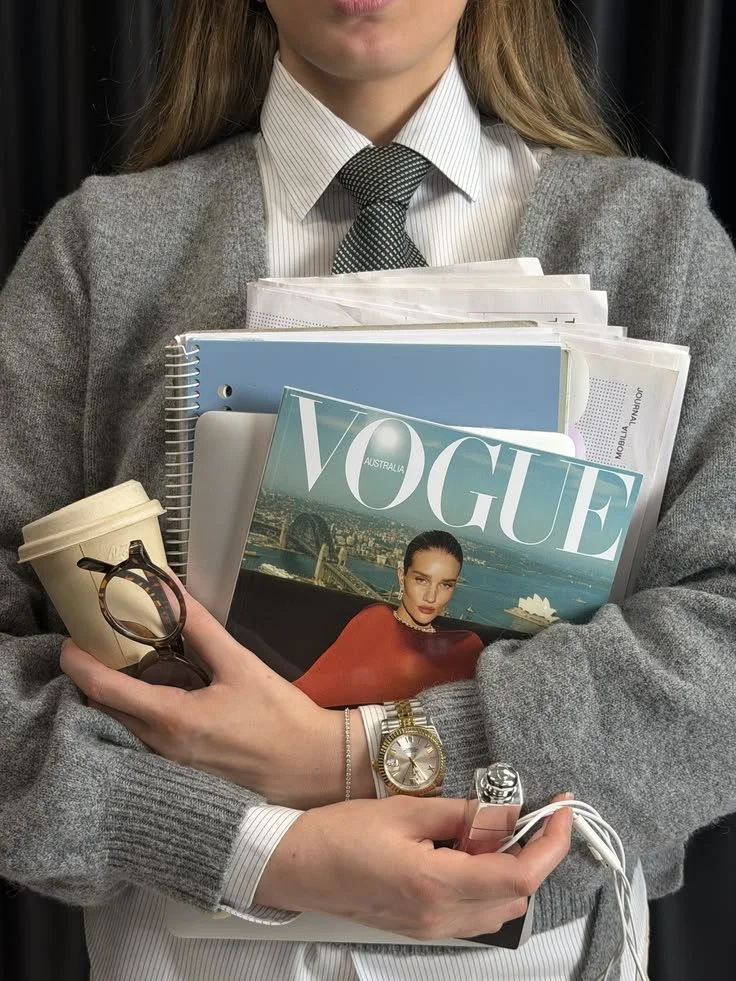 Person wearing a gray sweater and white dress shirt holding a coffee cup, sunglasses, a Vogue magazine, notebooks, and electronic devices.