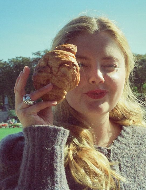 Close-up of a woman with blonde hair inside a car, making a funny face with red lipstick, holding a large croissant close to her face, with a sunny outdoor background.