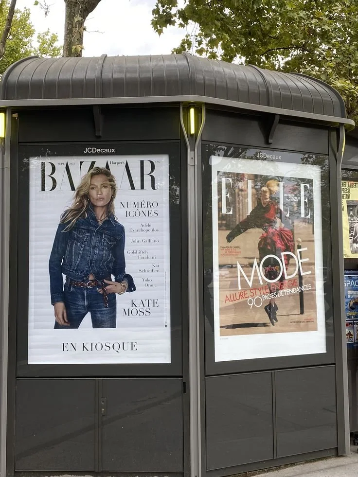 Two magazine advertisements in a black kiosk, one for Harper's Bazaar featuring Kate Moss in a denim jacket and jeans, and another for Elle magazine showcasing a woman in stylish clothing.