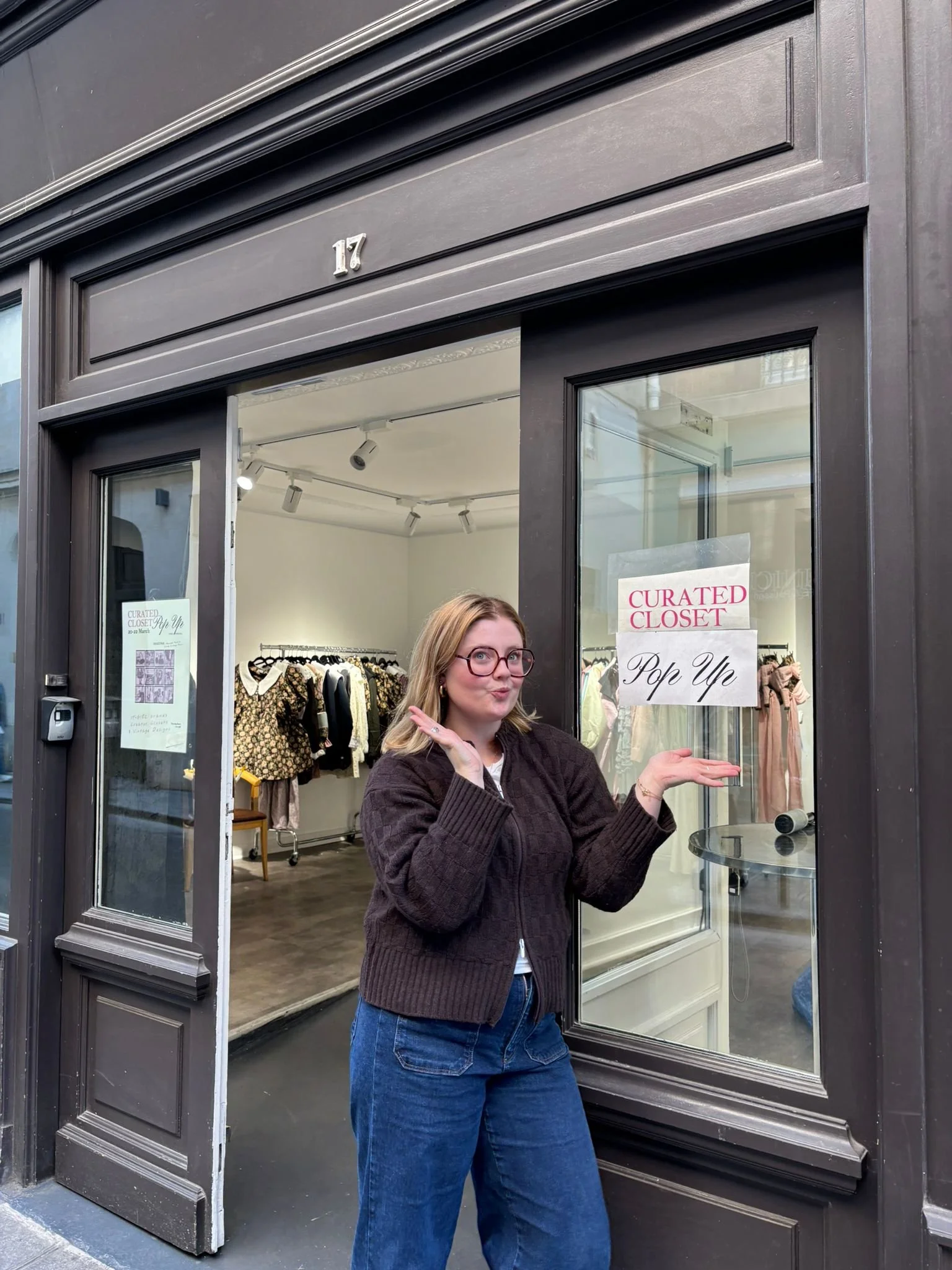 A woman standing in front of a store window with a playful gesture, wearing glasses, a dark brown sweater, and blue jeans, with a clothing store inside visible through the glass.