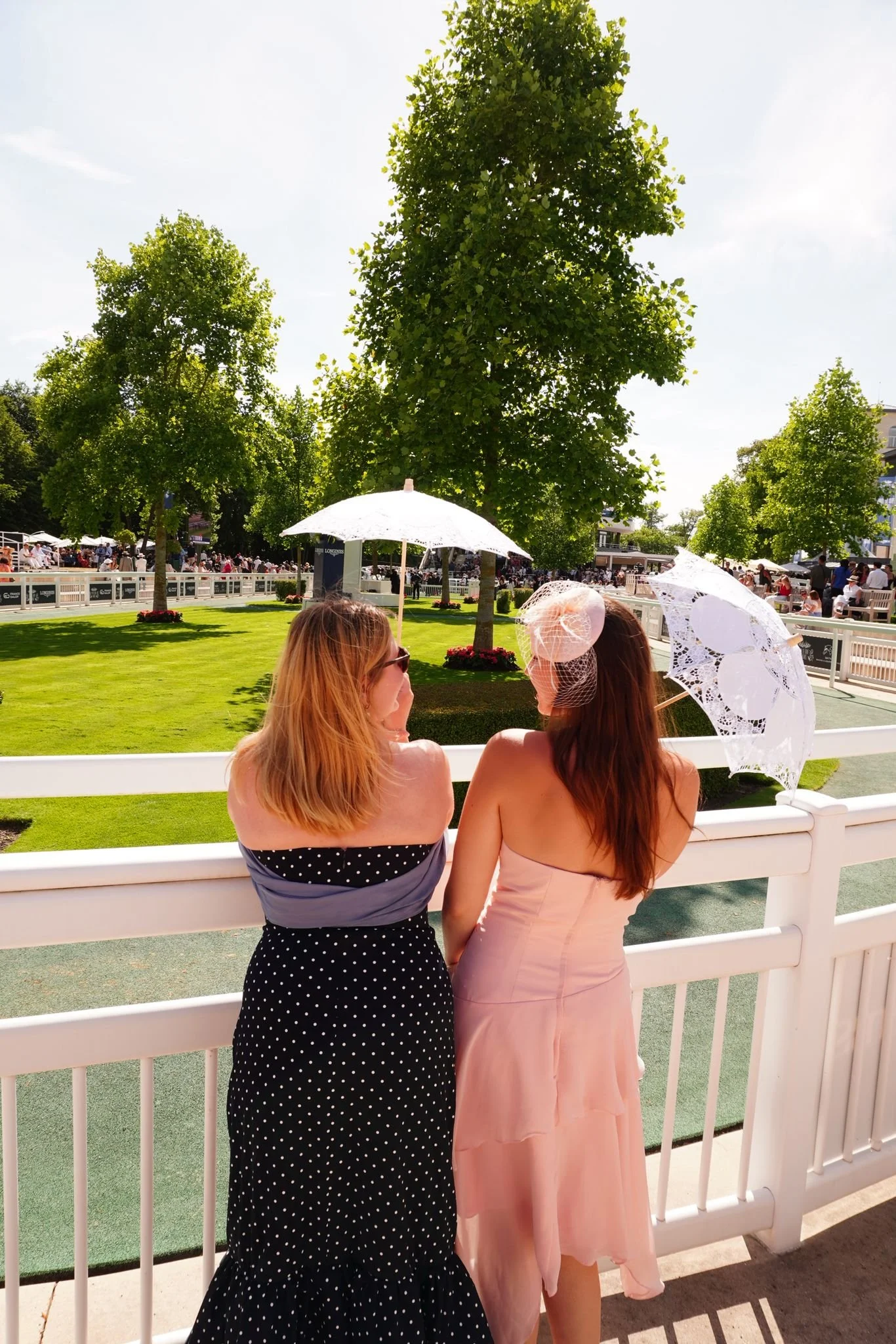 Two women in elegant dresses standing on a bridge during the day, holding lacy parasols, with a large grassy area, trees, and a crowd in the background.