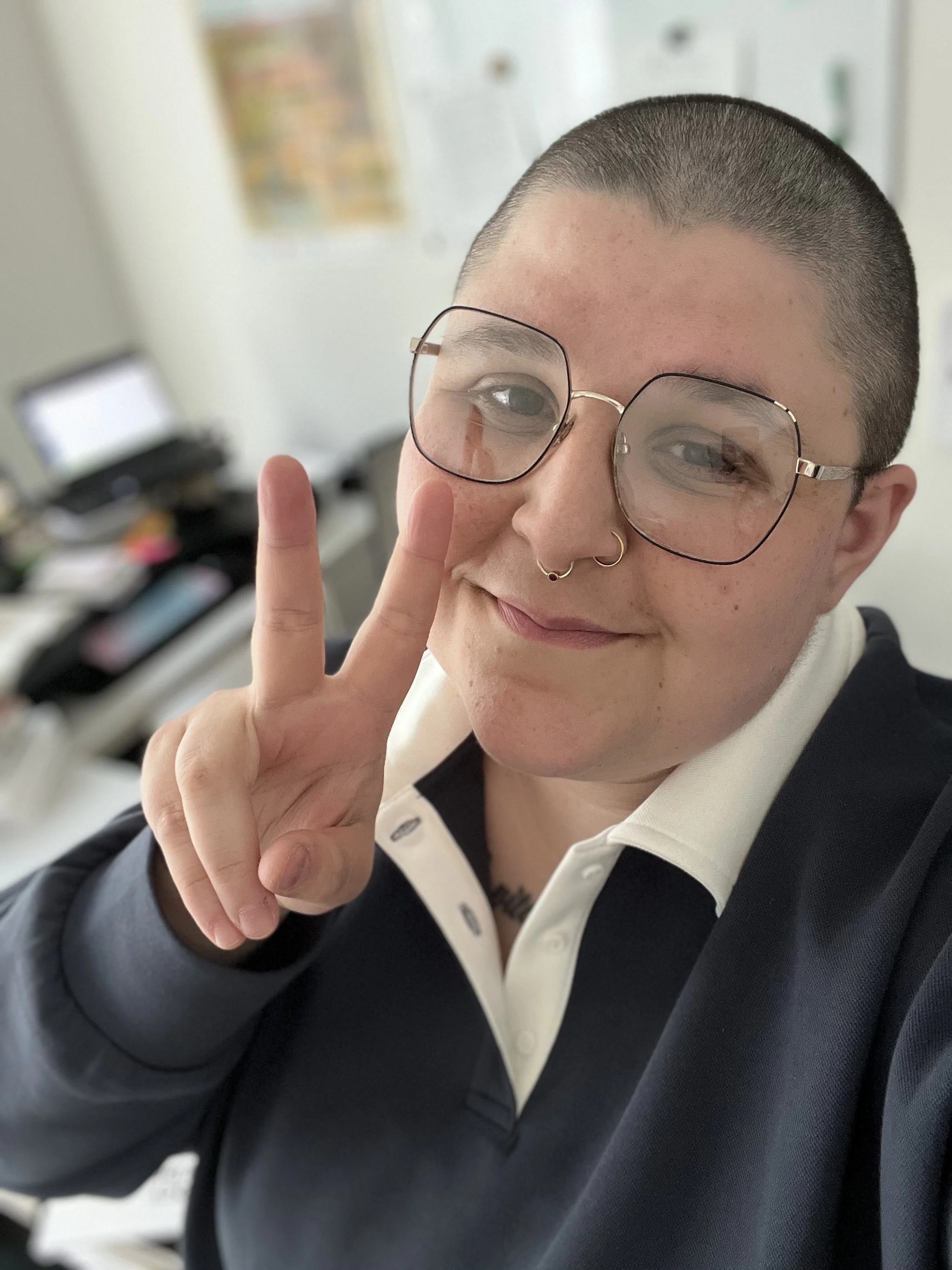 A person with short hair, wearing glasses and a septum piercing, making a peace sign with their hand, and smiling.