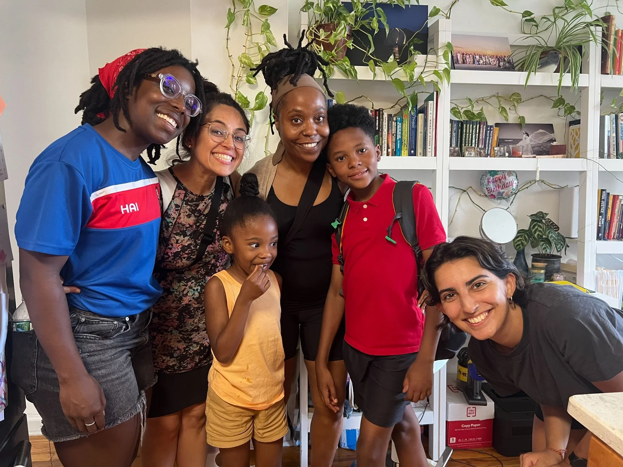 Group of five people smiling and posing together in a room with bookshelves and plants in the background.