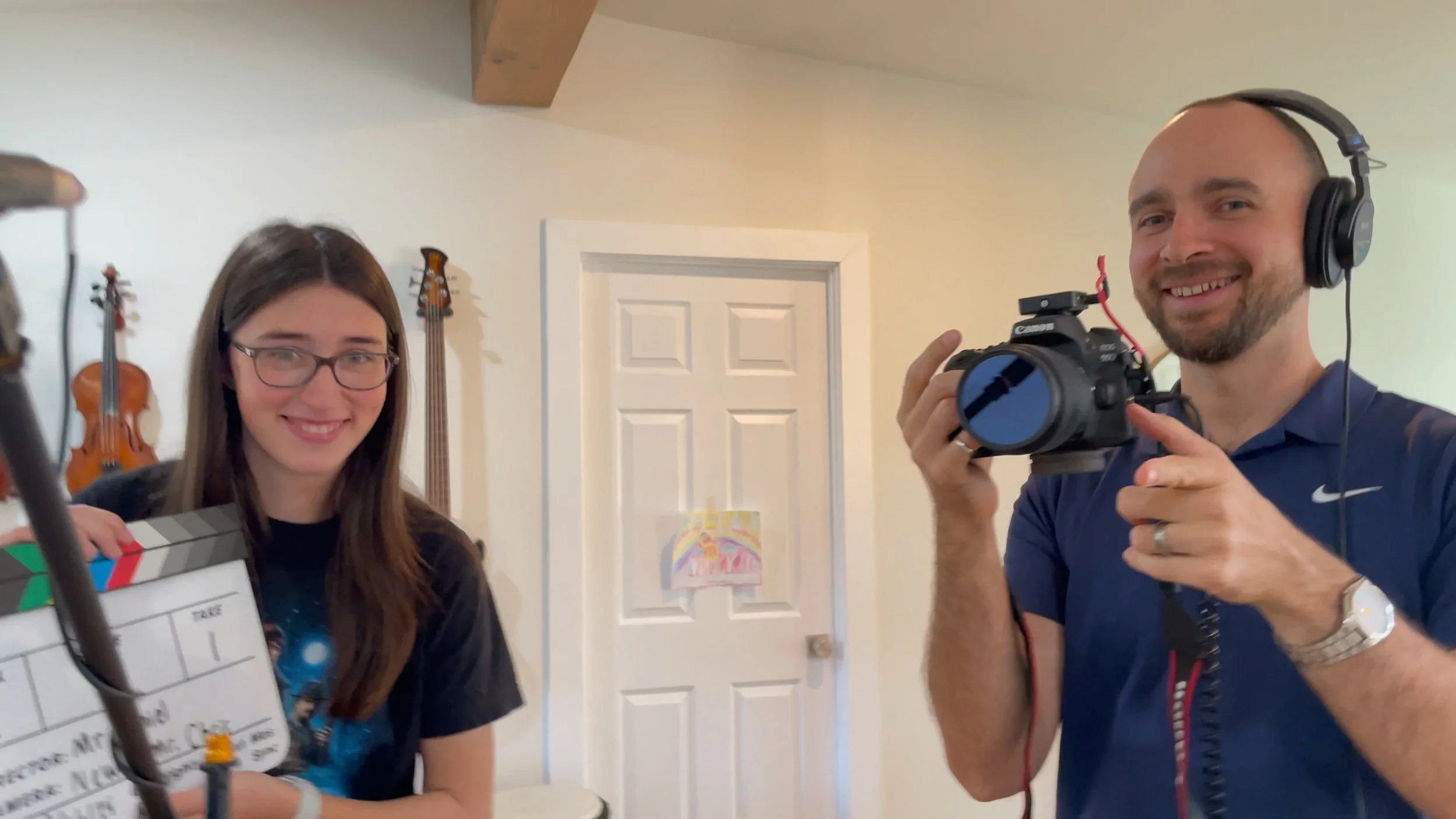 A young woman holding a clapperboard and a man with a camera wearing headphones smiling in a room with musical instruments on the wall.