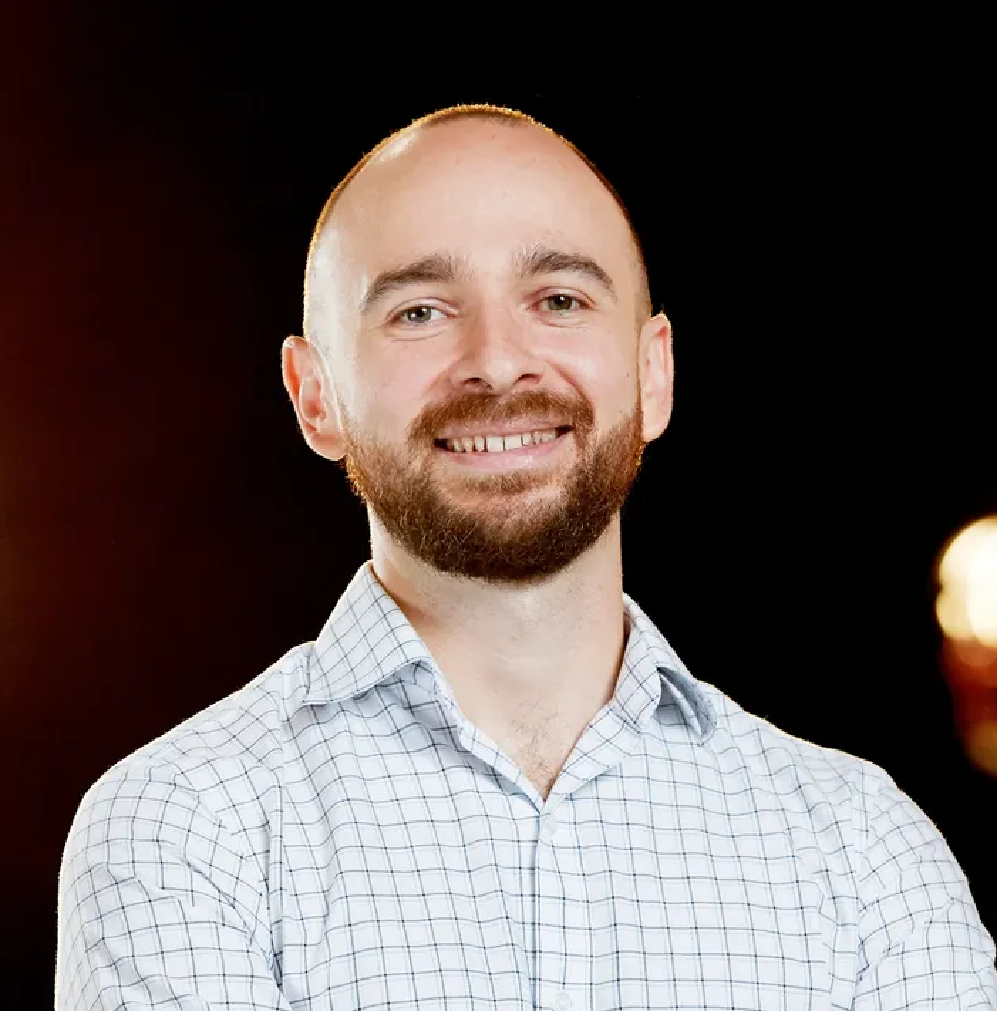 A smiling man with a beard and short hair, wearing a light checkered shirt, standing against a dark background with blurred warm lights.