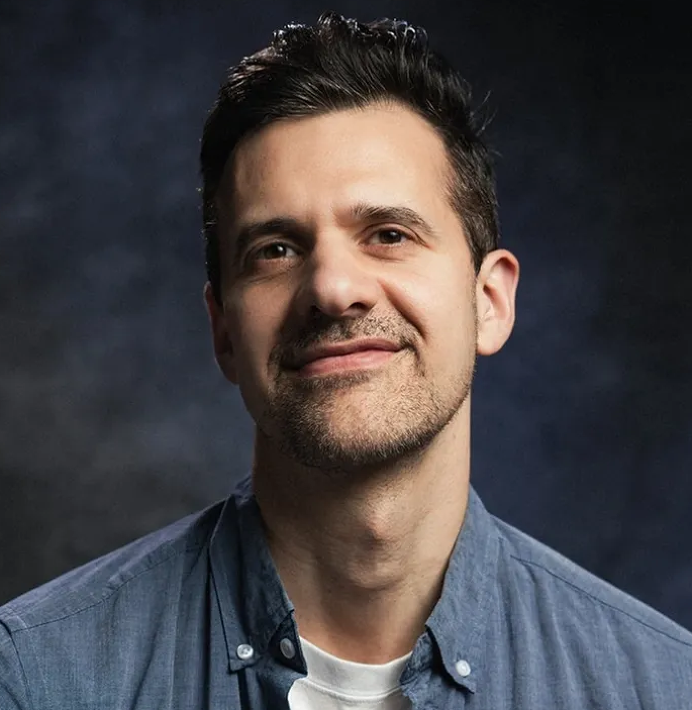 Headshot of a man with dark hair, slight beard, wearing a blue shirt and a white undershirt, in front of a dark background.