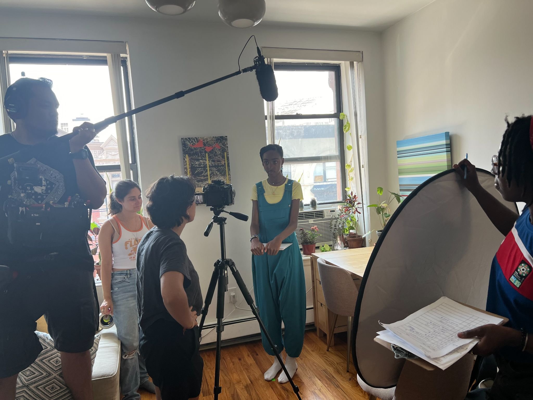 People involved in a film or video production inside a room with windows, including a person on the left holding a boom microphone, a person with a camera on a tripod, and a young woman standing in front of the camera. Others are seated or standing nearby, with a person on the right holding papers and a reflector, while a crew member is seen in the foreground.