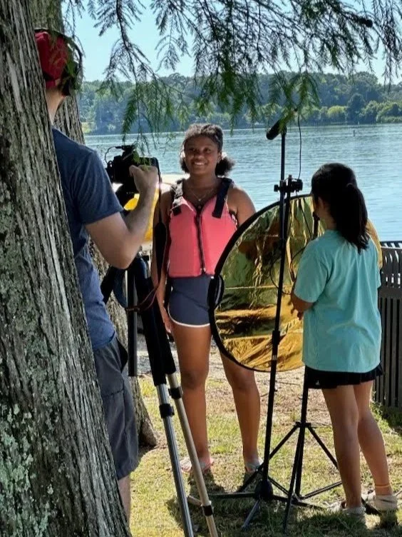 Two young girls appear to be filming a portrait by a lake with a camera and lighting equipment, surrounded by trees and nature.