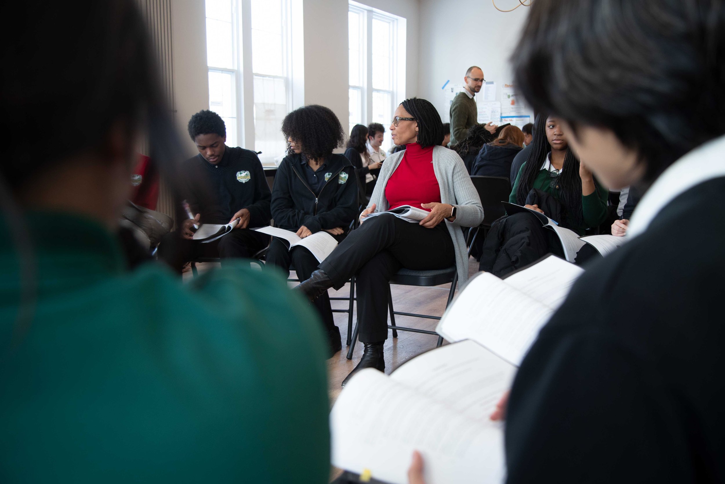 A diverse group of people seated in a classroom or conference room, some reading and others listening attentively. A woman in a red top and glasses is sitting with a notebook, while other individuals are engaged with papers. A man in a green sweater stands at the back near a bulletin board with papers, taking notes. The room has large windows letting in natural light.