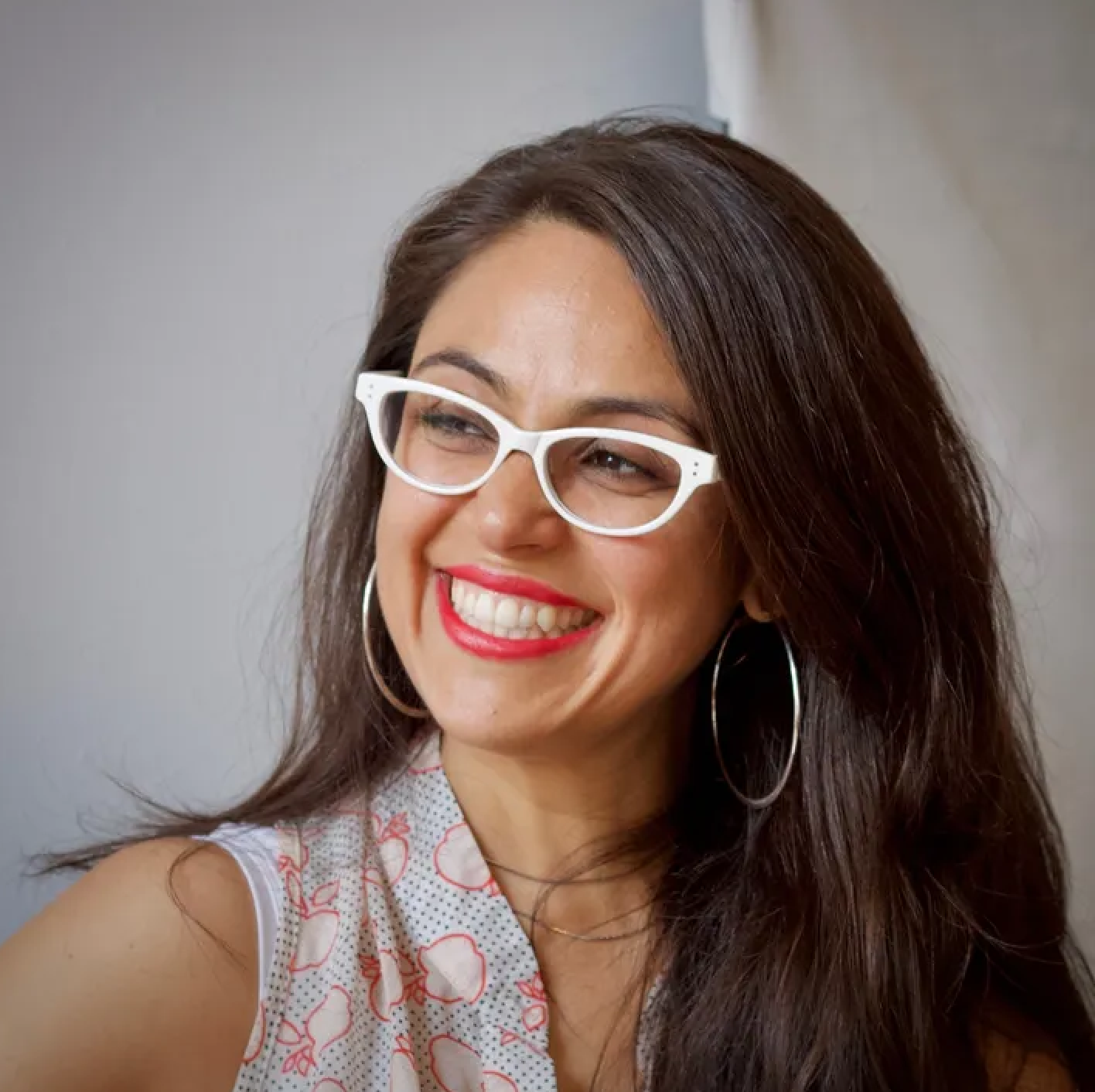 A woman smiling with white glasses, red lipstick, and hoop earrings, wearing a sleeveless top with a red and white pattern, with shoulder-length dark hair and a gray background.