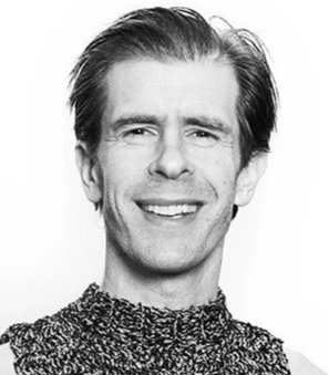 Sort-hvid portrætfoto af en smilende mand med mørkt, pjusket hår, iført en tekstureret sweater.