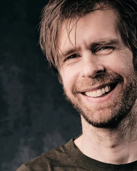 Mand med skæg, der smiler bredt, iført en sort T-shirt, mod en mørk baggrund.