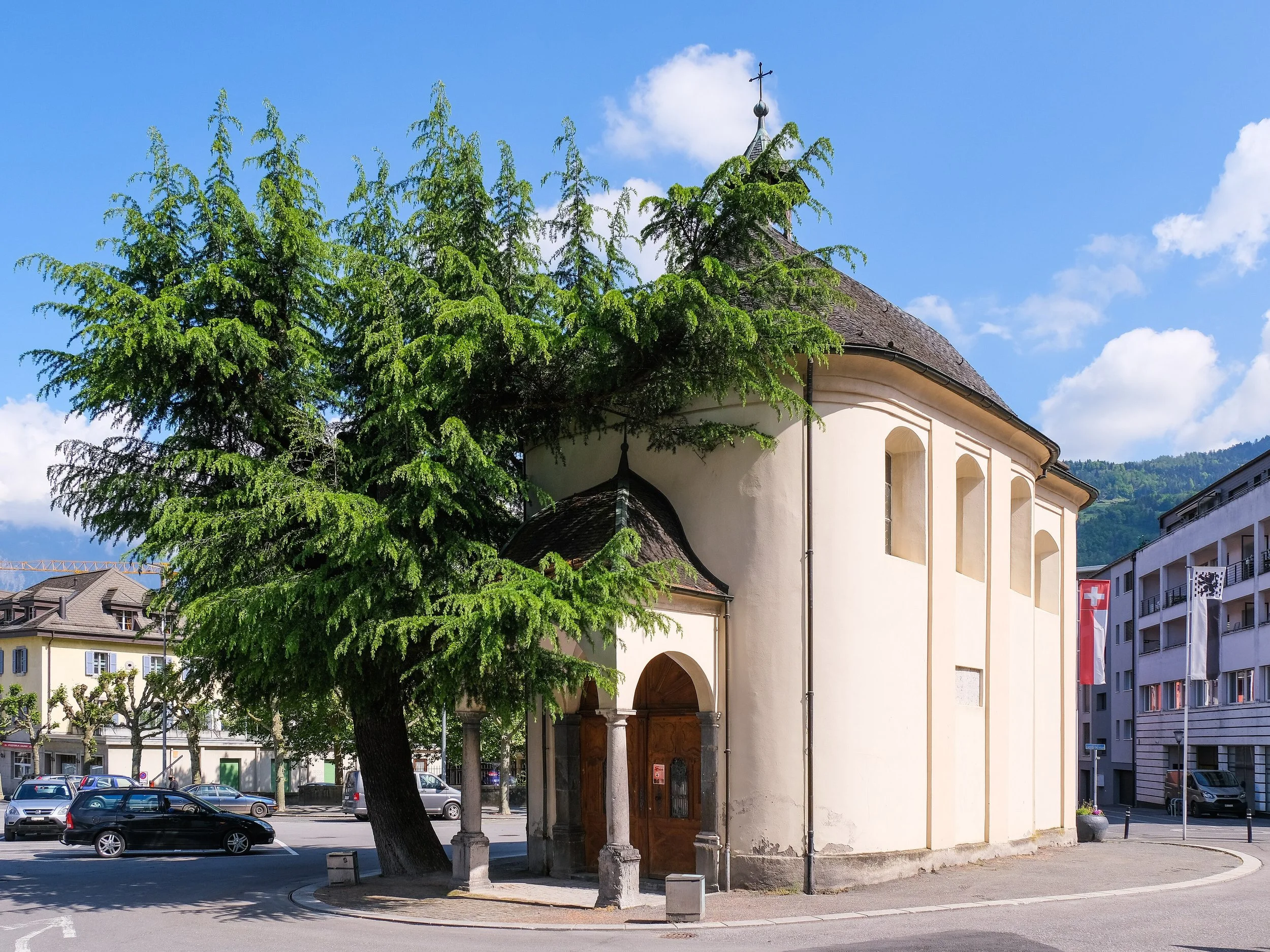 Une petite église en pierre blanche avec un toit en ardoise, entourée d'arbres et située en milieu urbain, sous un ciel bleu avec quelques nuages.