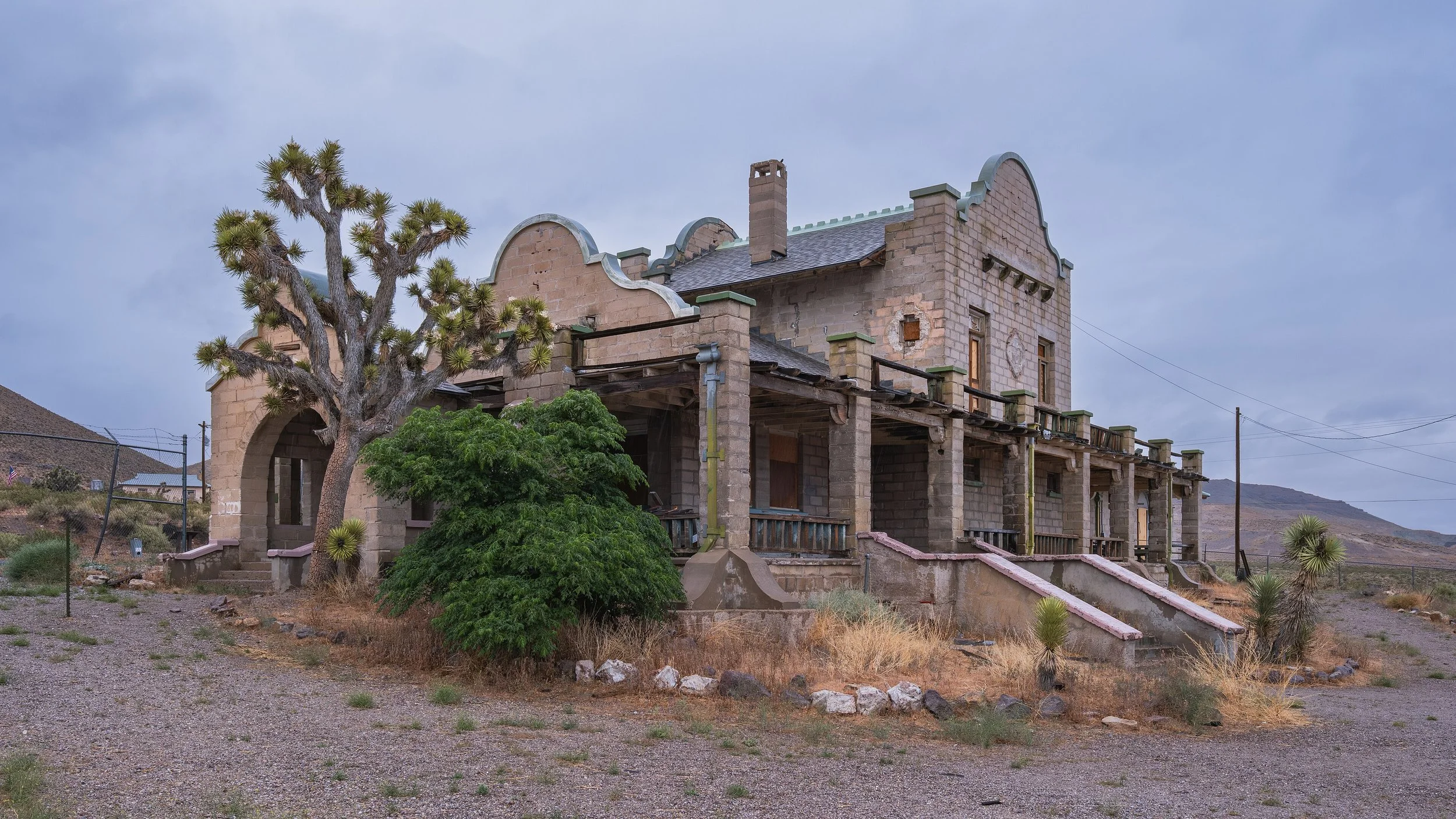 Une maison en pierre inachevée avec un balcon, des fenêtres sans vitres, située dans un paysage désertique, avec des cactus et des broussailles autour.
