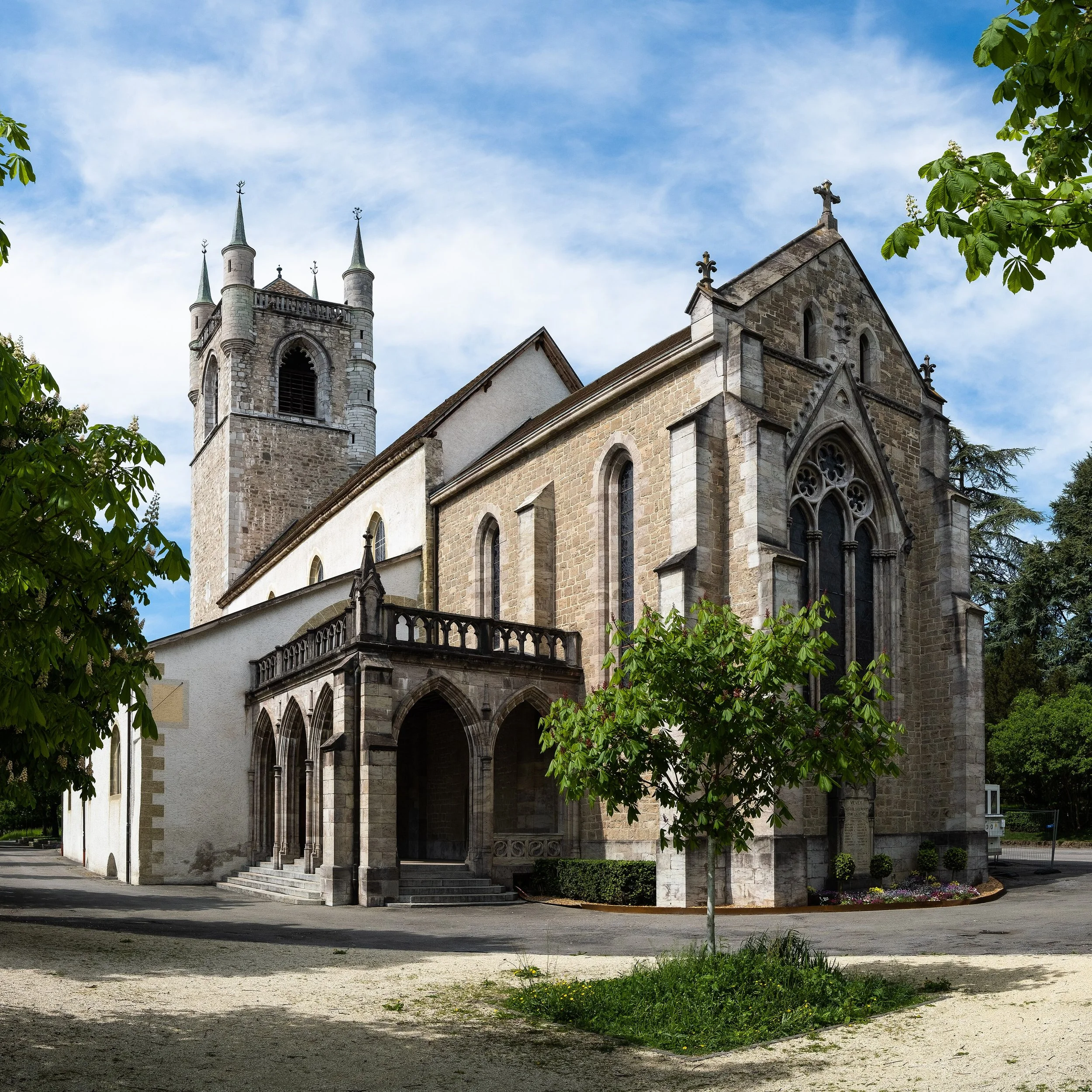 Vue d'une ancienne église en pierre avec deux tours, un ciel bleu avec quelques nuages, entourée d'arbres verts.