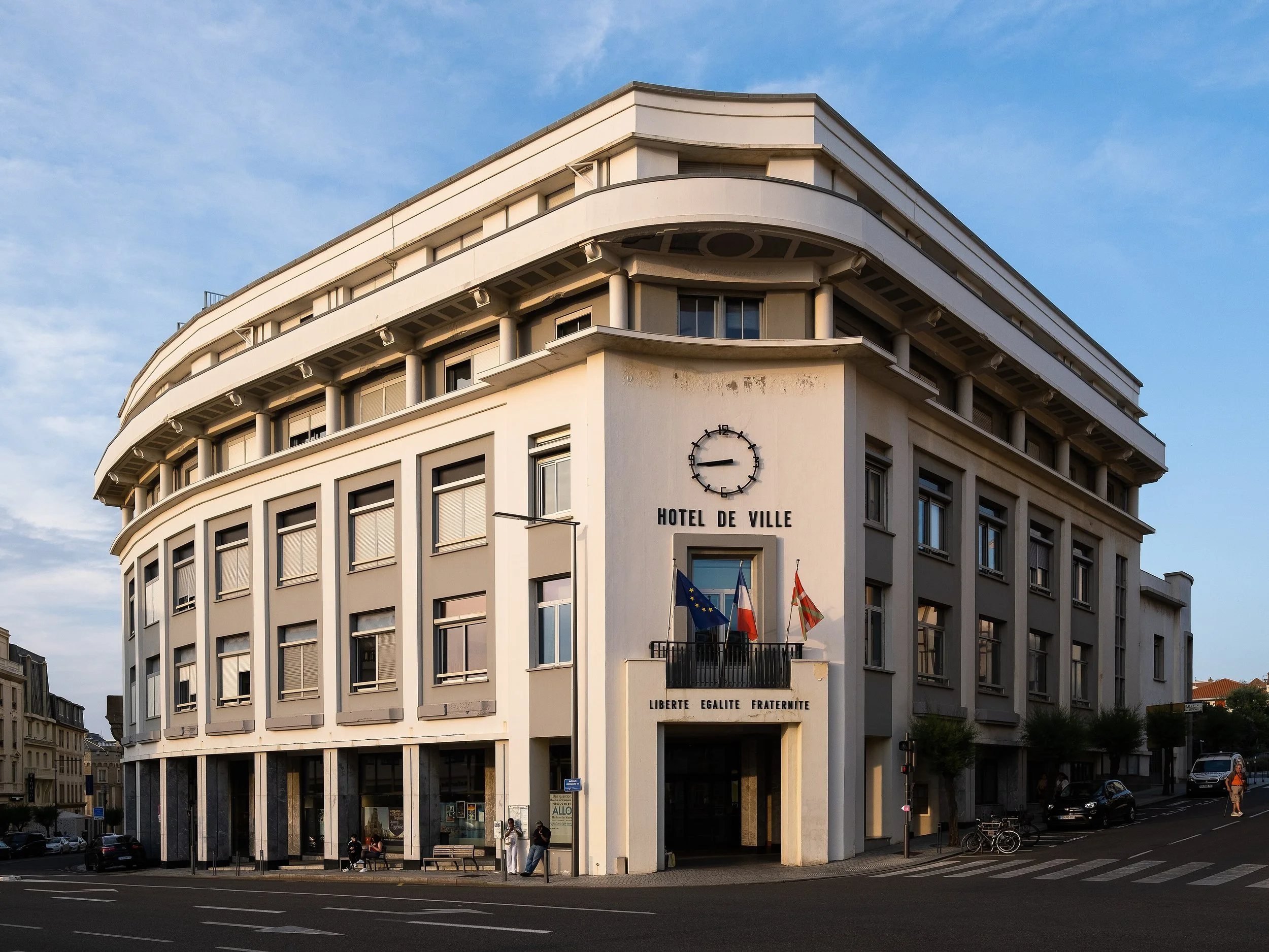 Bâtiment de style art déco avec des drapeaux français, européens et régionaux, une horloge et une phrase en français sur la façade, situé dans une rue avec des voitures et des personnes.