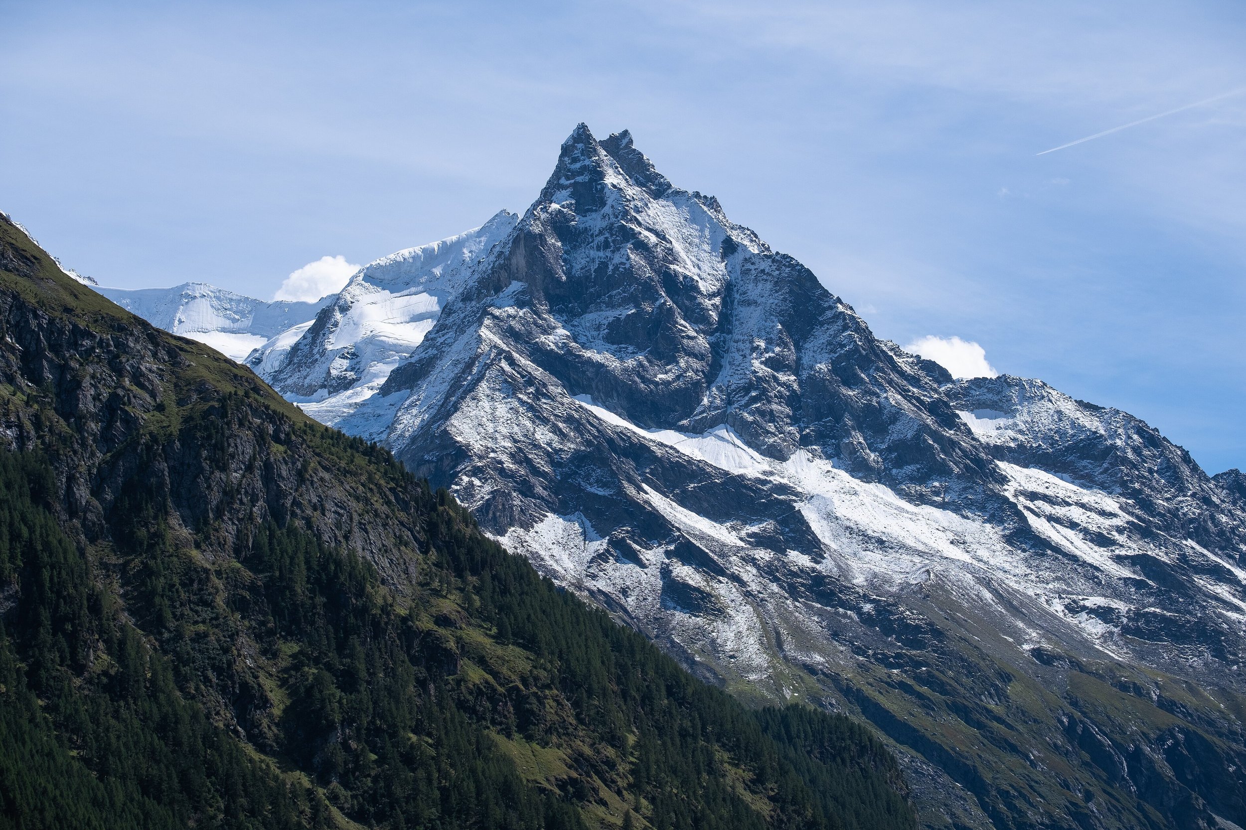 Une montagne enneigée avec un sommet pointu et des pentes rocheuses, entourée de forêts vertes en bas.