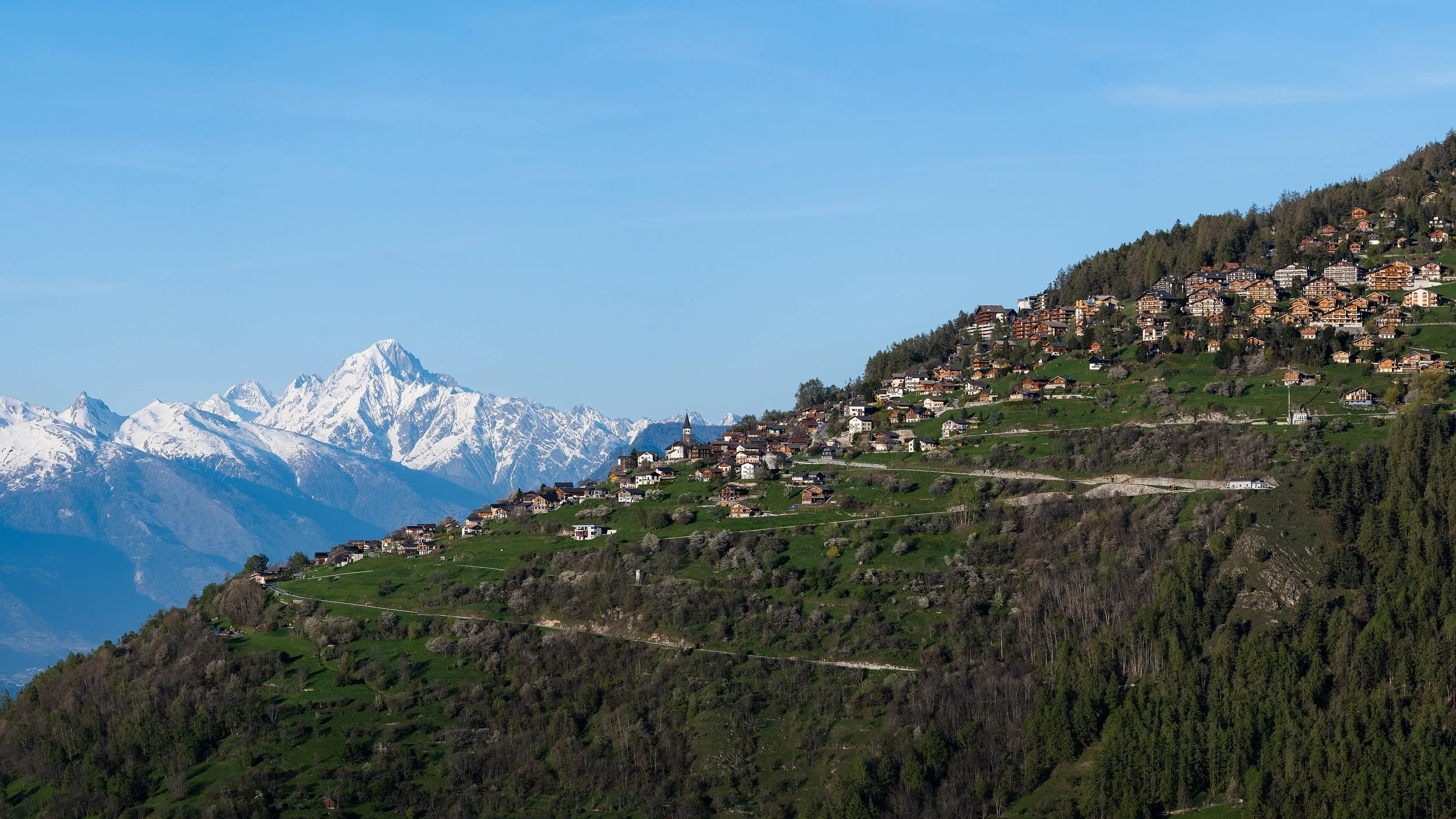 Village de montagne avec des maisons en bois sur une colline verdoyante, avec des montagnes enneigées à l'arrière-plan et un ciel bleu clair.