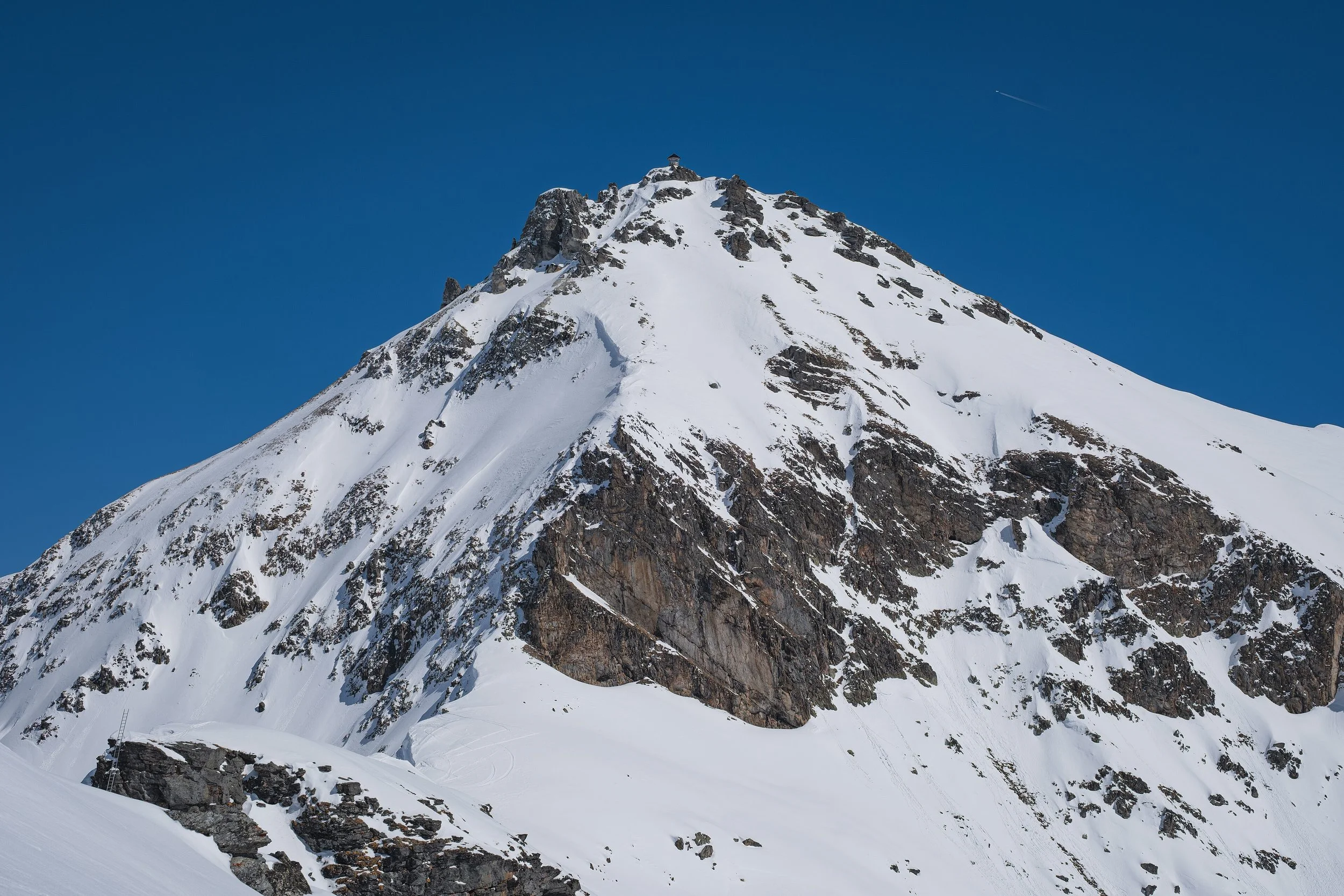 Montagne enneigée avec un sommet rocheux et un bâtiment en haut, ciel bleu clair, peu de nuages.