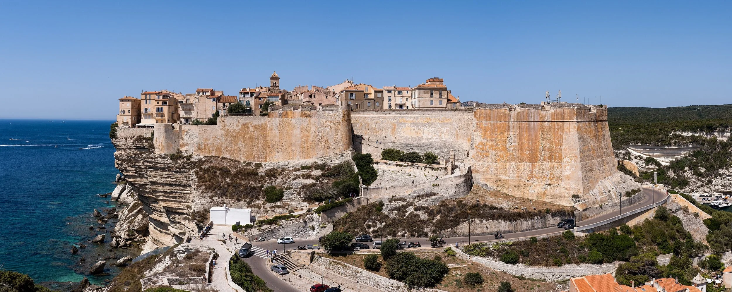 Vue panoramique d'une ville médiévale perchée sur une falaise calcaire surplombant la mer Méditerranée avec des rues étroites, des bâtiments en pierre et des murailles anciennes.