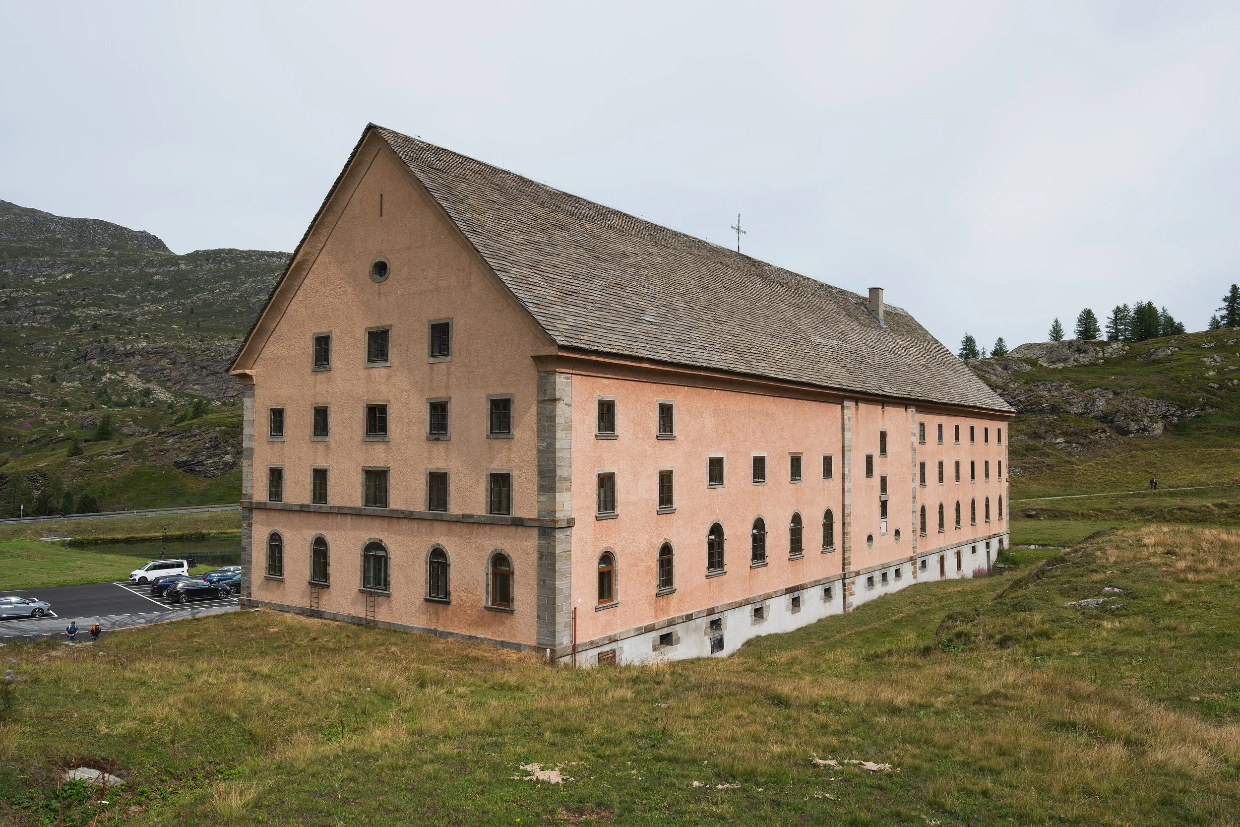 Immeuble de plusieurs étages avec fenêtres, situé dans un paysage de montagnes verdoyantes, parking avec voitures devant, ciel nuageux.