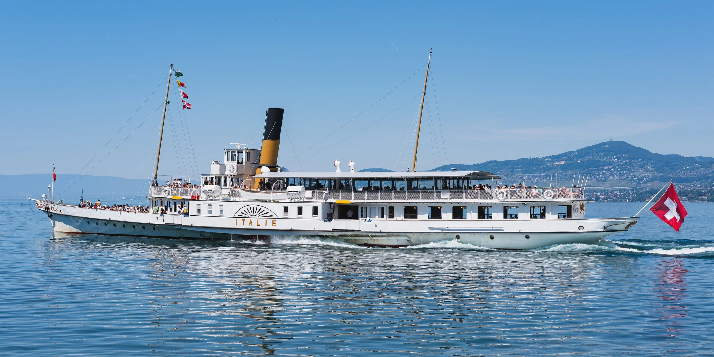 Un bateau à vapeur blanc portant le nom 'Italie' naviguant sur un lac avec des passagers à bord. Drapeau suisse au derrière et drapeaux en haut des mâts, avec un paysage montagneux en arrière-plan.